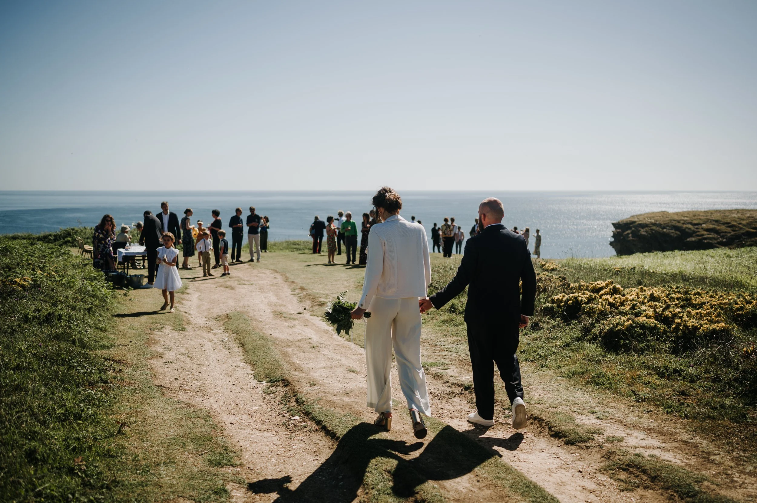 Un couple se tenant la main lors d'un mariage en plein air près de la mer, avec des invités en arrière-plan.