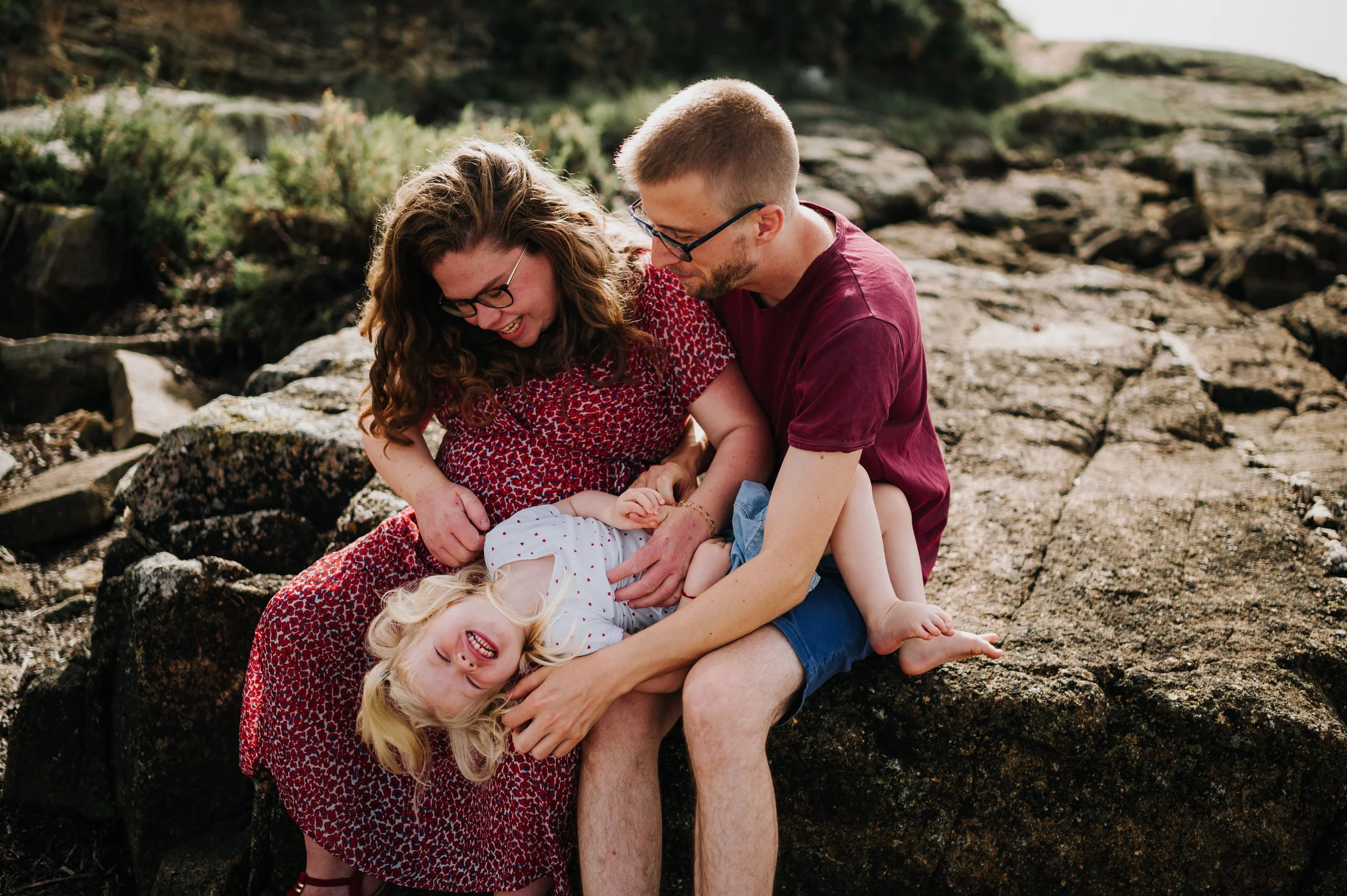 Une famille de quatre personnes, deux adultes et deux enfants, souriants et riant, assis sur des rochers en plein air, lors d'une journée ensoleillée.
