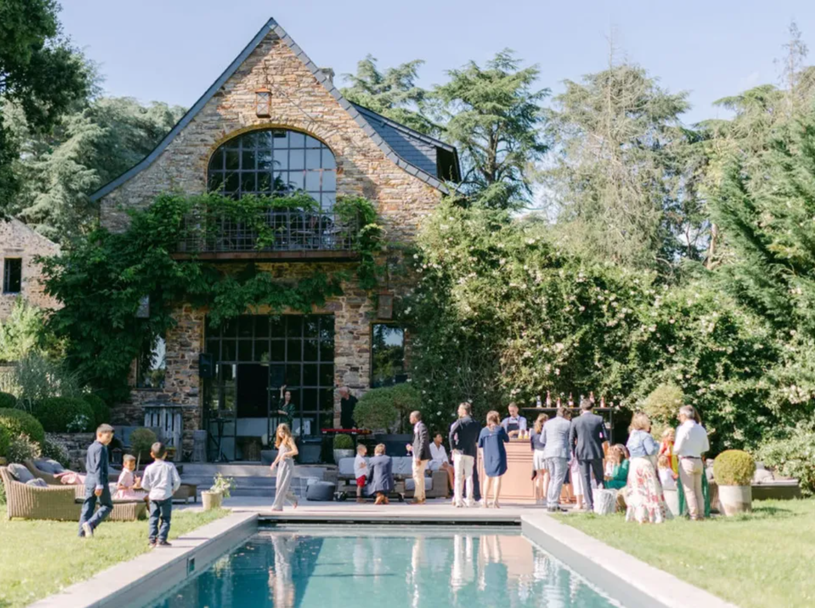 Une fête ou réception en extérieur devant une grande maison en pierre avec un jardin, des personnes habillées élégamment, une scène avec des musiciens, et une piscine en premier plan.
