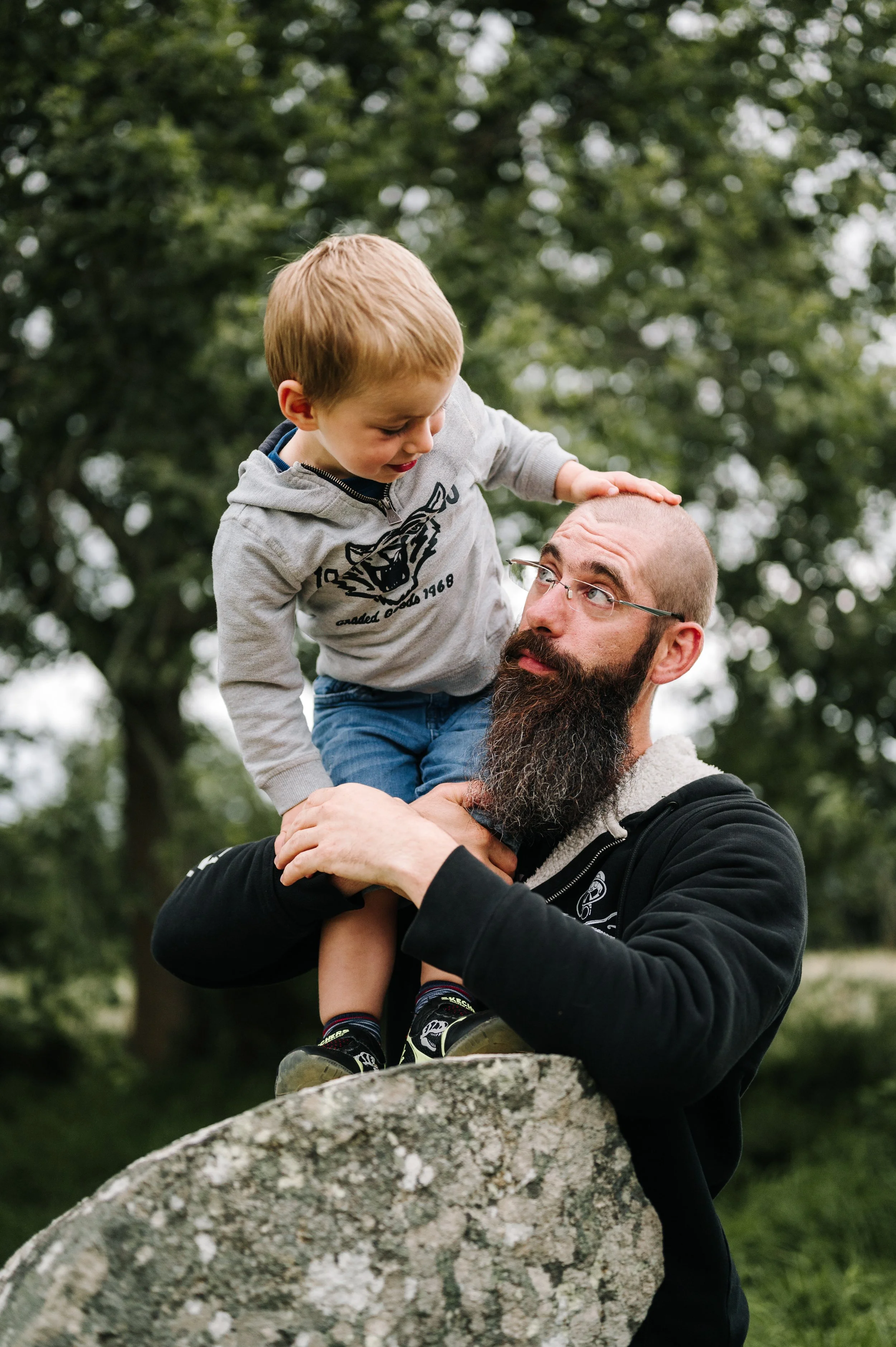 Un homme avec une barbe portant des lunettes, tenant un enfant blond sur ses épaules dans un parc, tous deux entourés d'arbres.