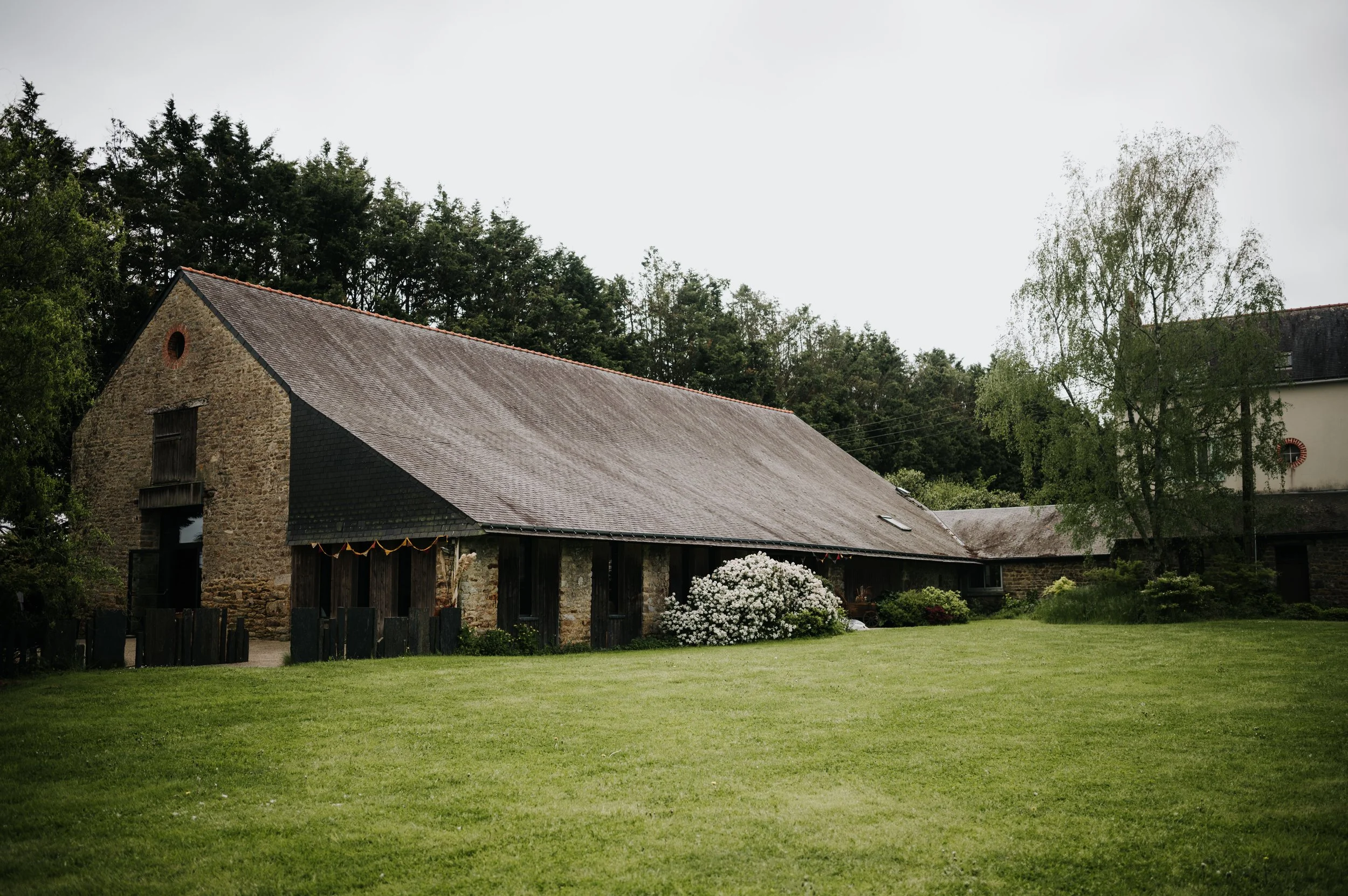 Une vieille ferme en pierre avec un toit en ardoise, entourée d'un grand jardin avec de l'herbe verte, des arbustes fleuris et des arbres en arrière-plan, sous un ciel gris.