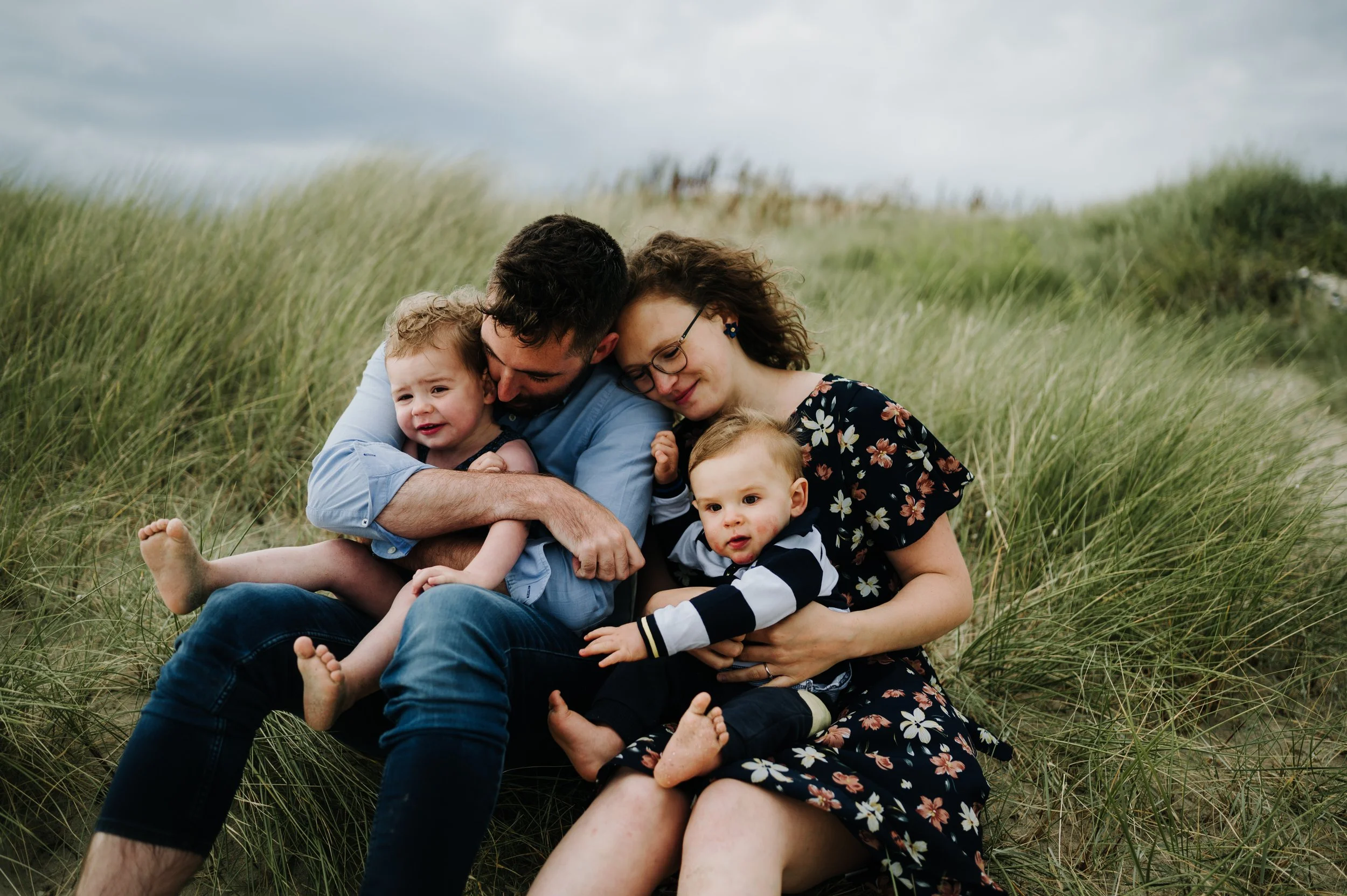 Une famille de quatre personnes, deux adultes et deux enfants, assis dans un champ d'herbes hautes, partageant un moment de tendresse et de bonheur en extérieur par une journée nuageuse.
