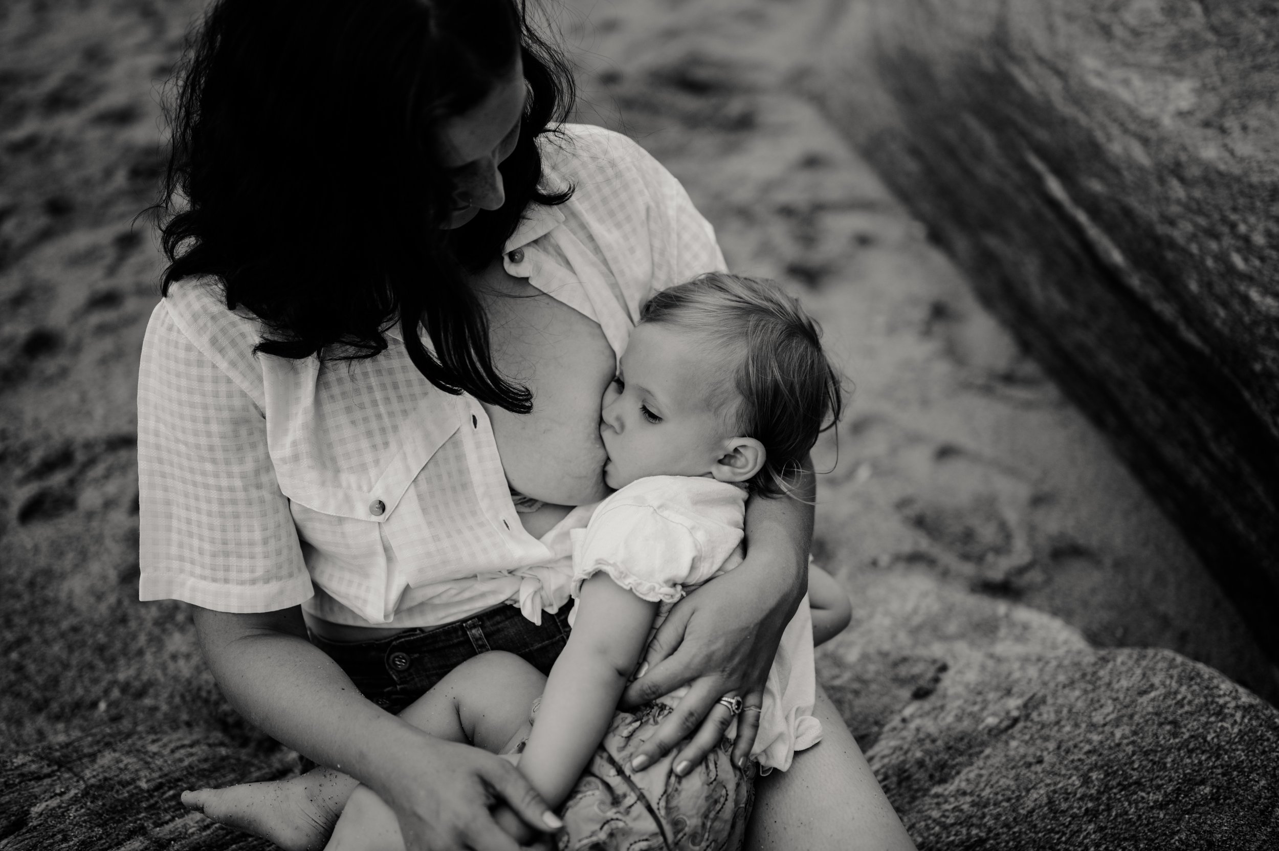 Une femme et un enfant sont en train de partager un moment affectueux à l'extérieur, probablement sur une plage ou un parc, avec un arbre en arrière-plan. La femme embrasse tendrement l'enfant sur la joue."