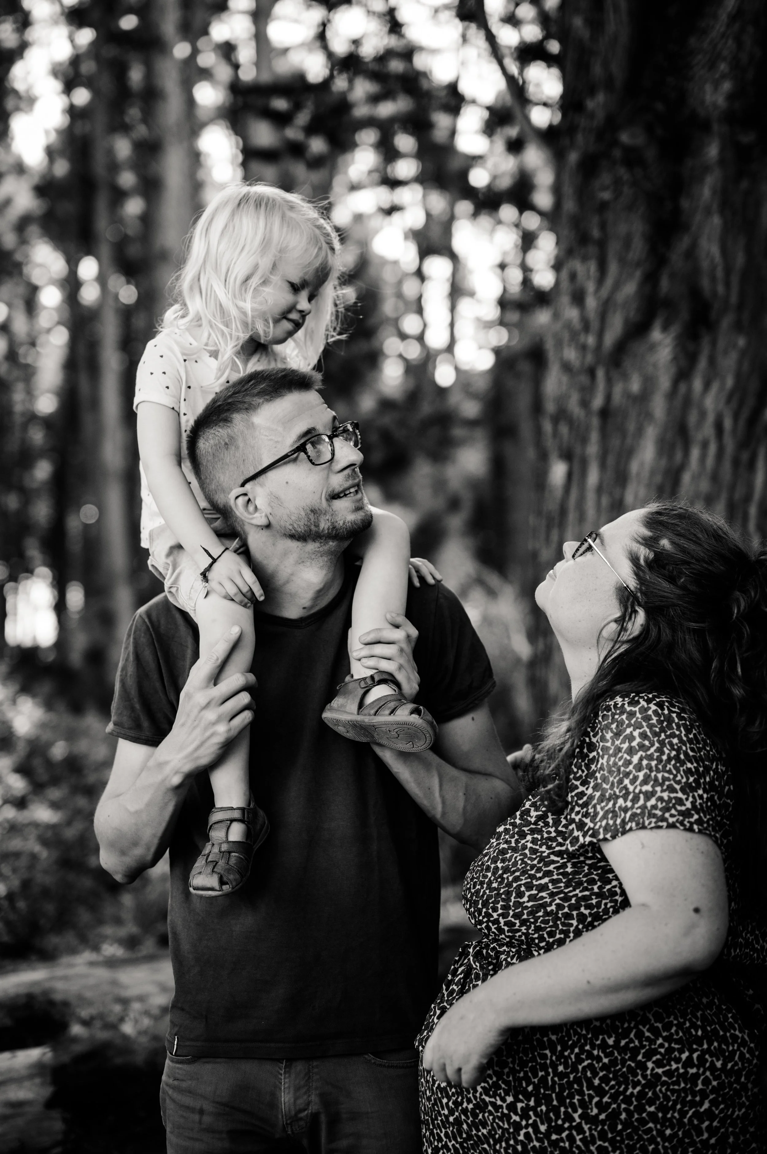 Une famille en forêt, un homme porte une petite fille sur ses épaules, une femme regarde la petite fille. L'image est en noir et blanc.