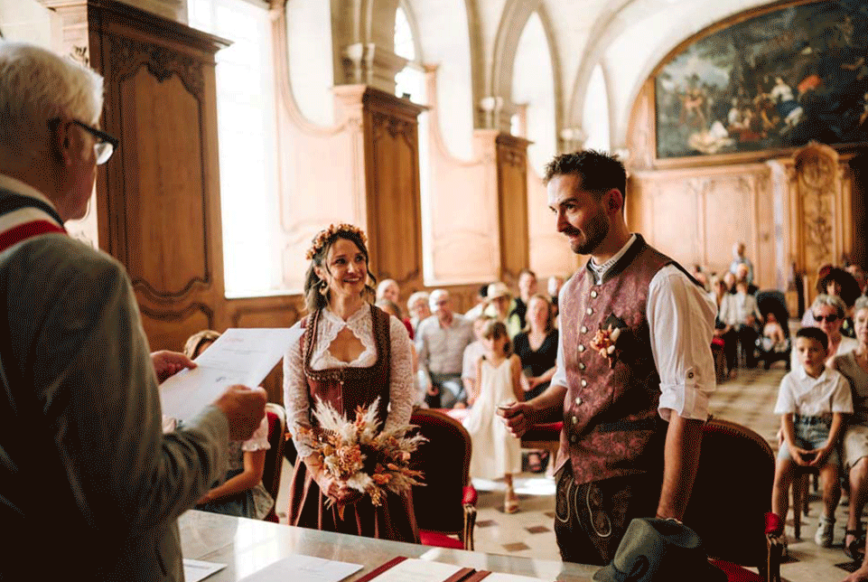 Un couple de mariage échange des vœux dans une salle ornée de bois, avec des invités en arrière-plan, lors d'une cérémonie nuptiale.