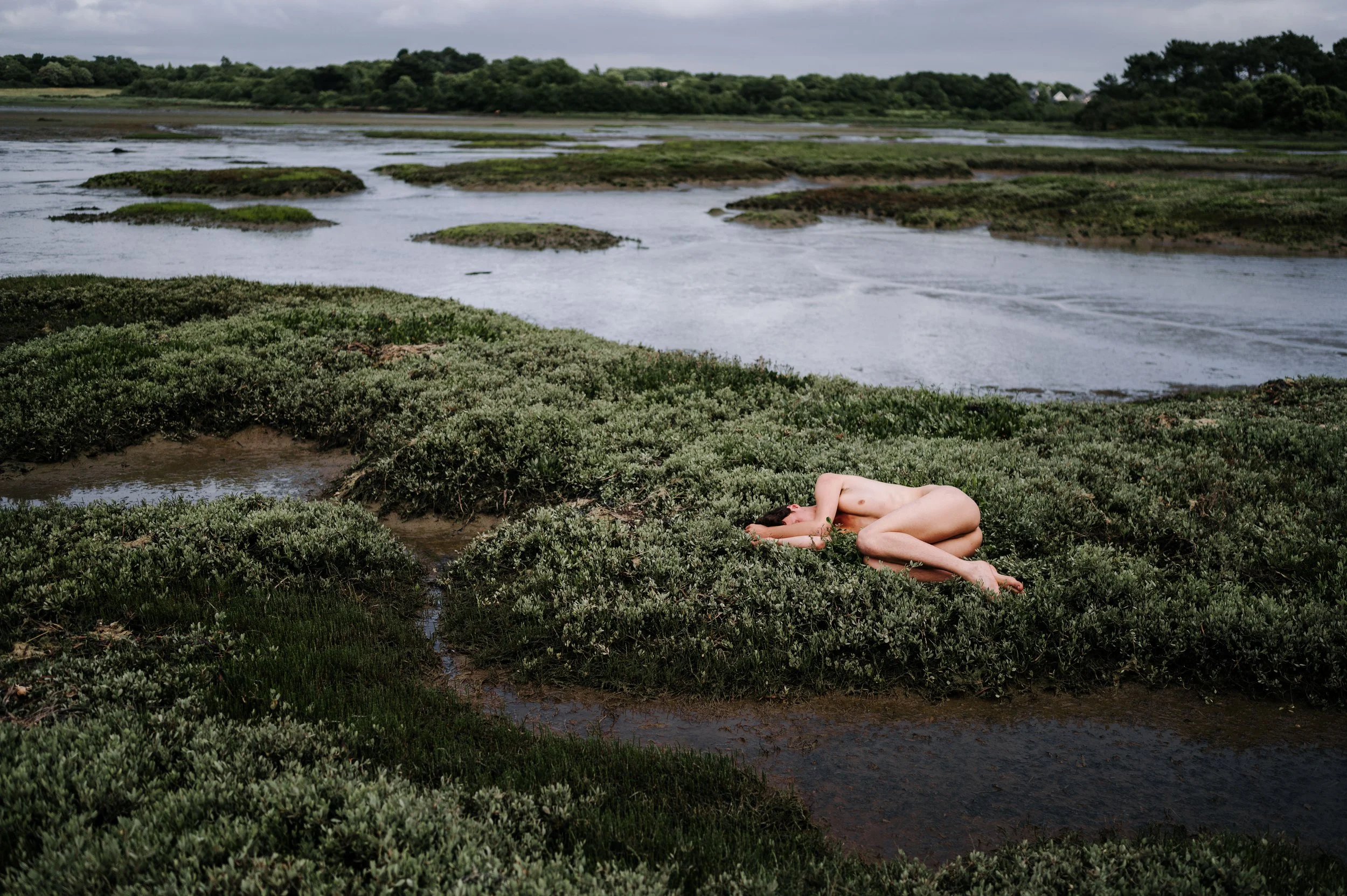 Une personne nue allongée dans une nature humide avec des plantes basses, près d'une rivière ou d'un étang, avec un ciel nuageux en arrière-plan.