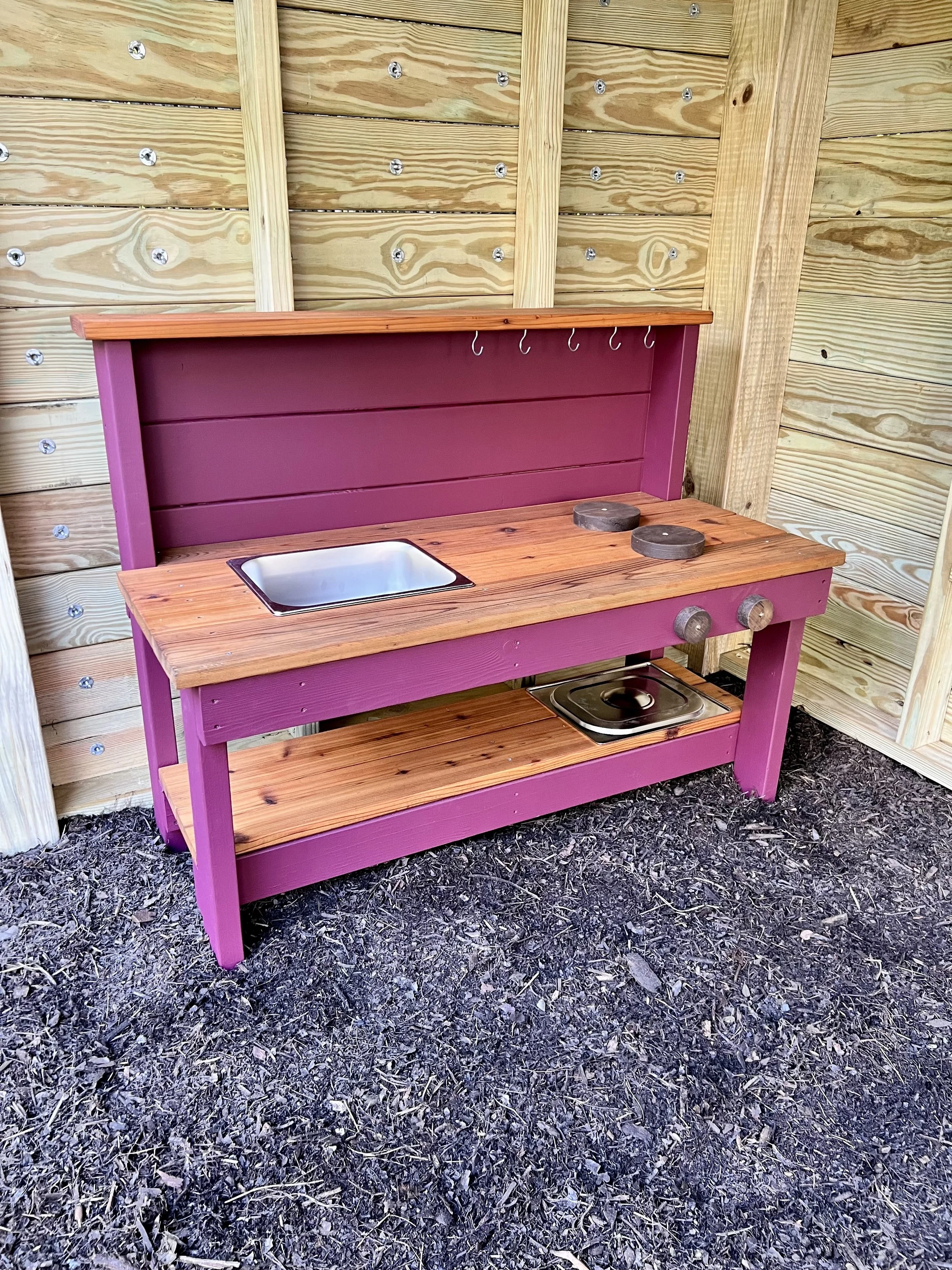 Pink wooden play kitchen with a small sink, two stovetop burners, and hooks for hanging items, situated outdoors on a dirt ground with a wooden fence background.