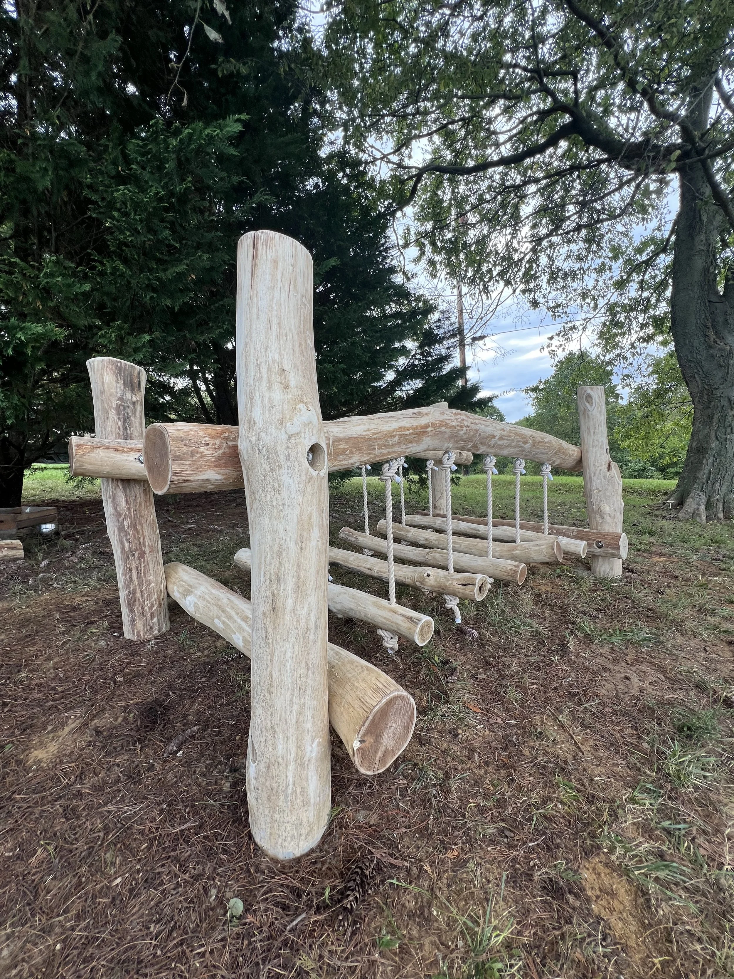 Wooden outdoor playground structure with swings and climbing elements, surrounded by grass, trees, and cloudy sky.