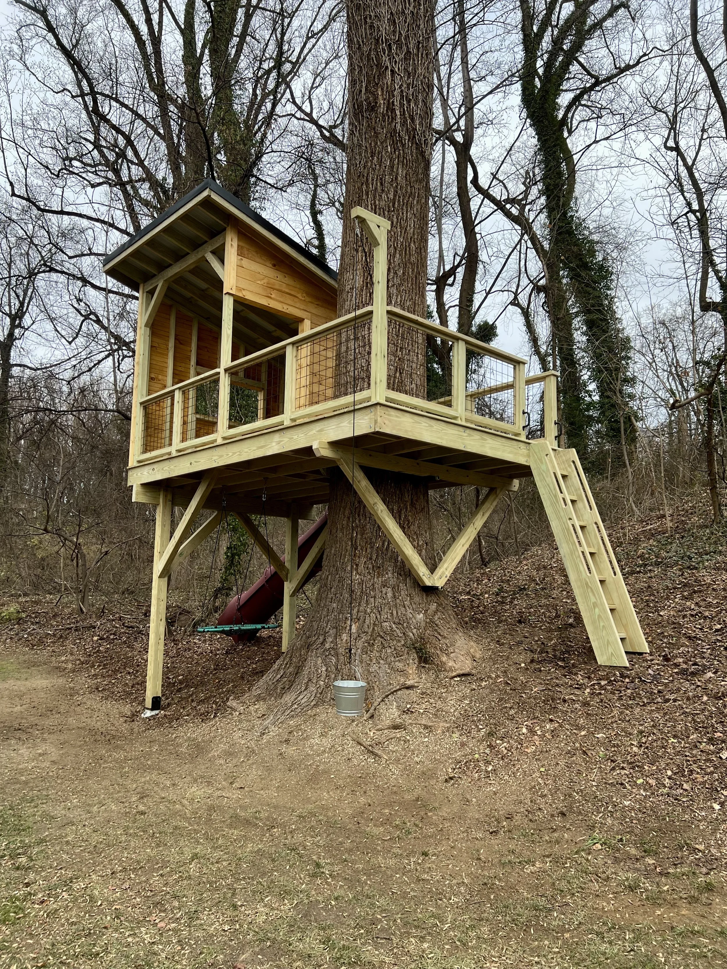 A treehouse built on a large tree in a wooded area with a wooden deck, railing, and a slide, and a bucket hanging from a string attached to the tree.