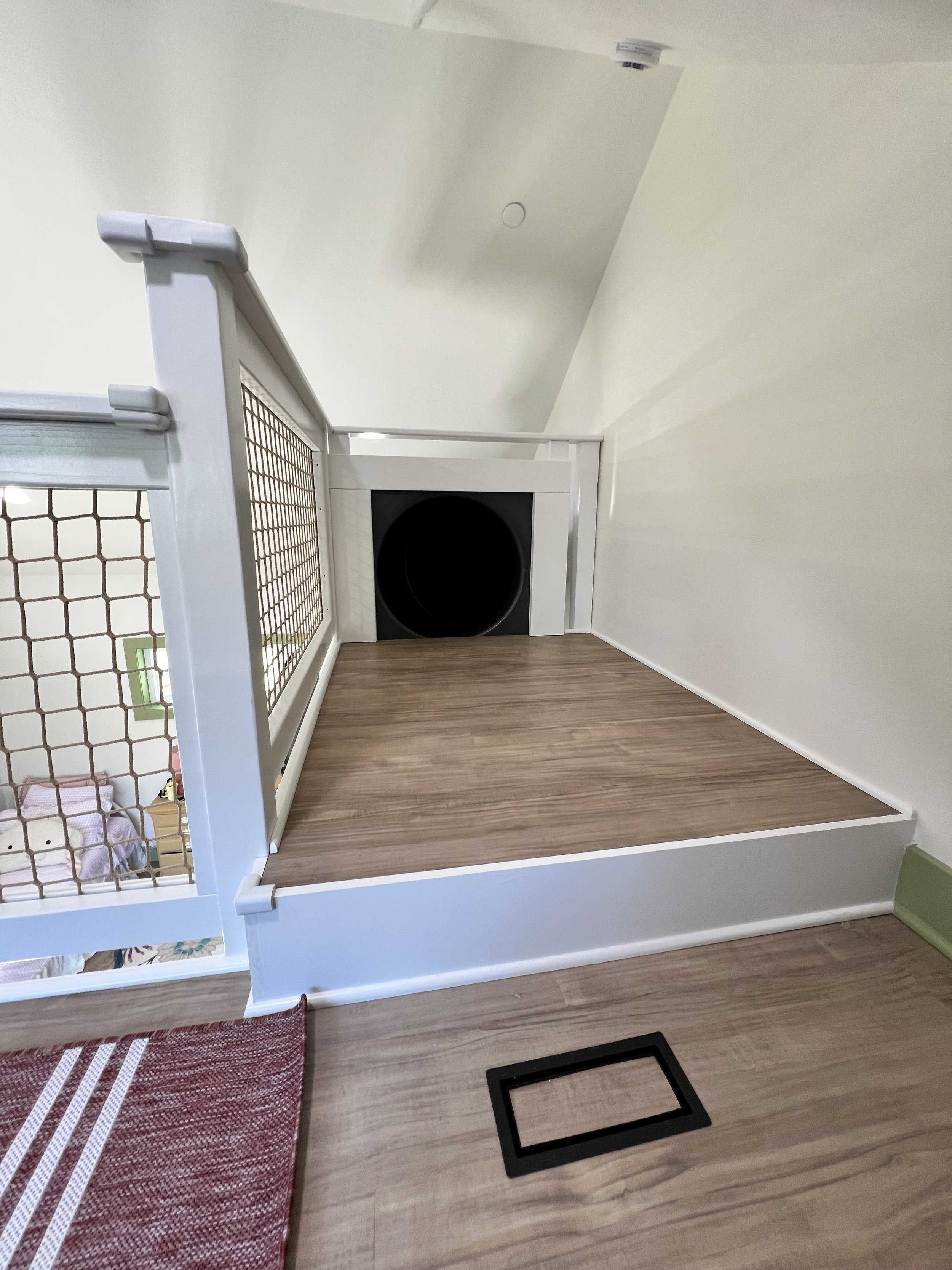 Close-up of a bedroom with a red and white striped rug, view of a section of a wooden floor, a window with light streaming in, and part of a bed with pink bedding.