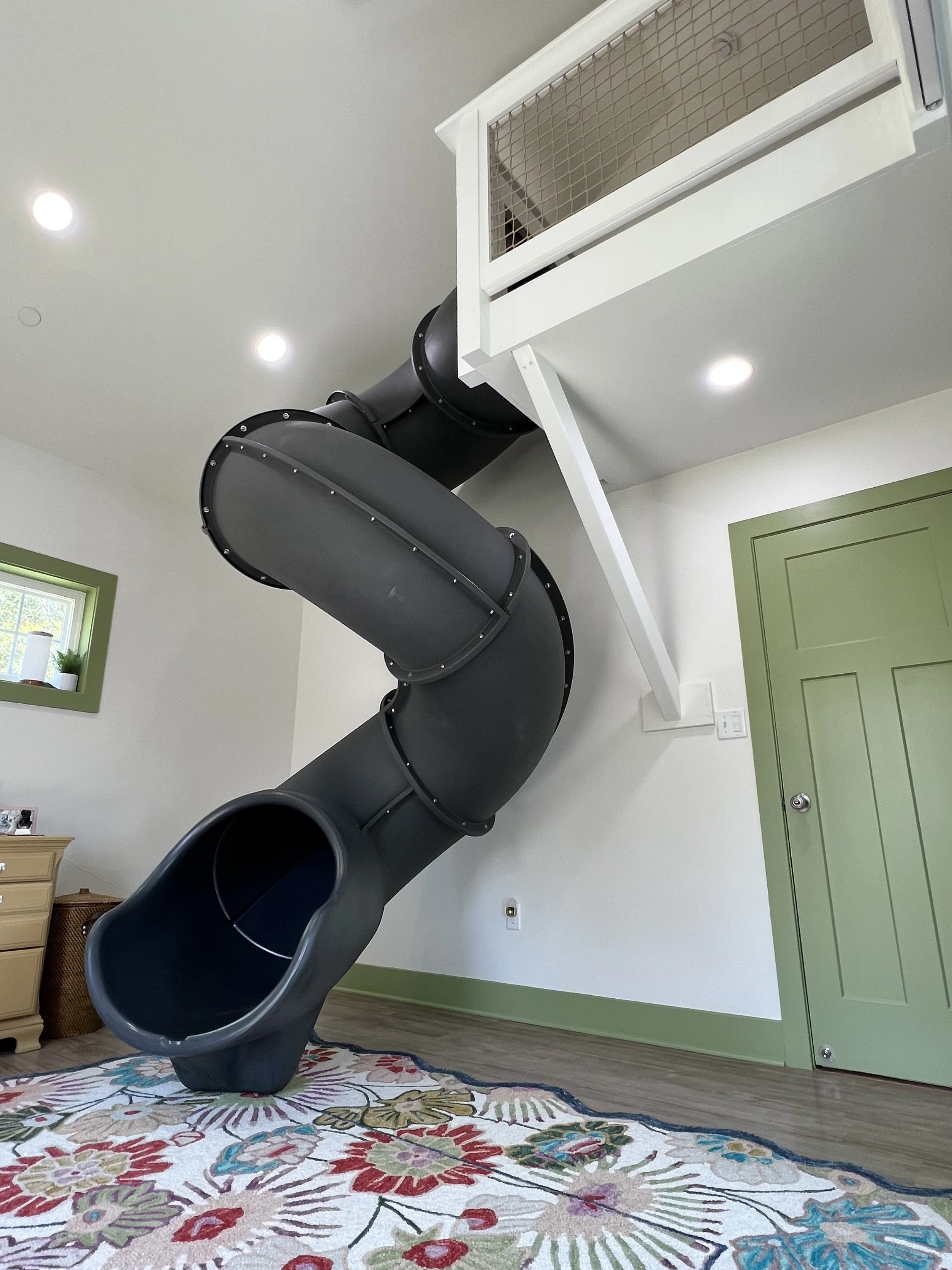 Indoors, there is a black spiral slide extending from a small second-floor landing with a white railing. The walls are white with green trim around a door and a window. The floor has a colorful floral-patterned rug. There are ceiling lights and a small dresser with picture frames and a plant.