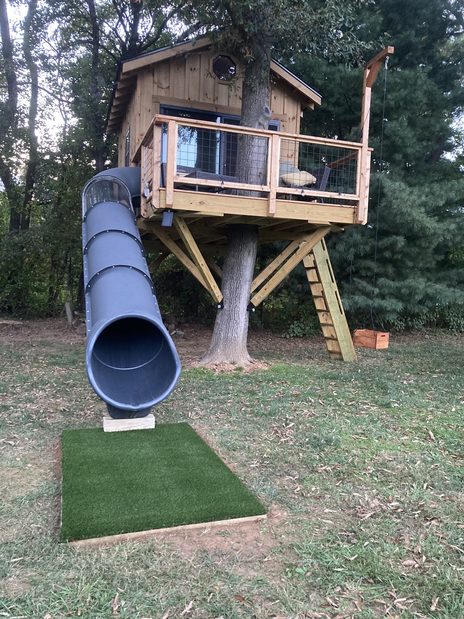 A treehouse built around a large tree, with a wooden deck porch, railing, and a metal slide attached to it, situated in a wooded yard with grass and fallen leaves.