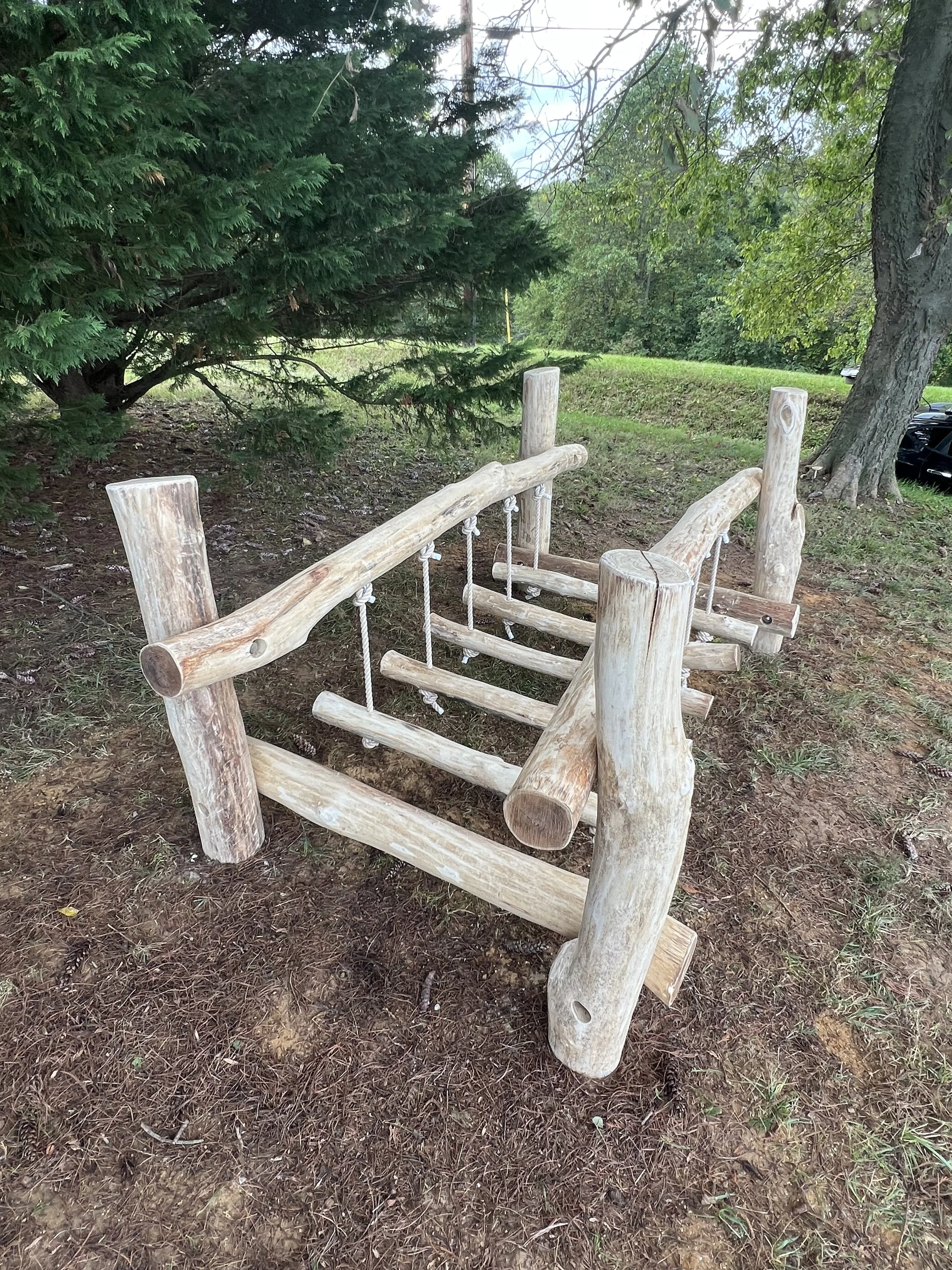 A wooden bench made from logs situated outdoors on grassy soil, surrounded by trees and greenery.