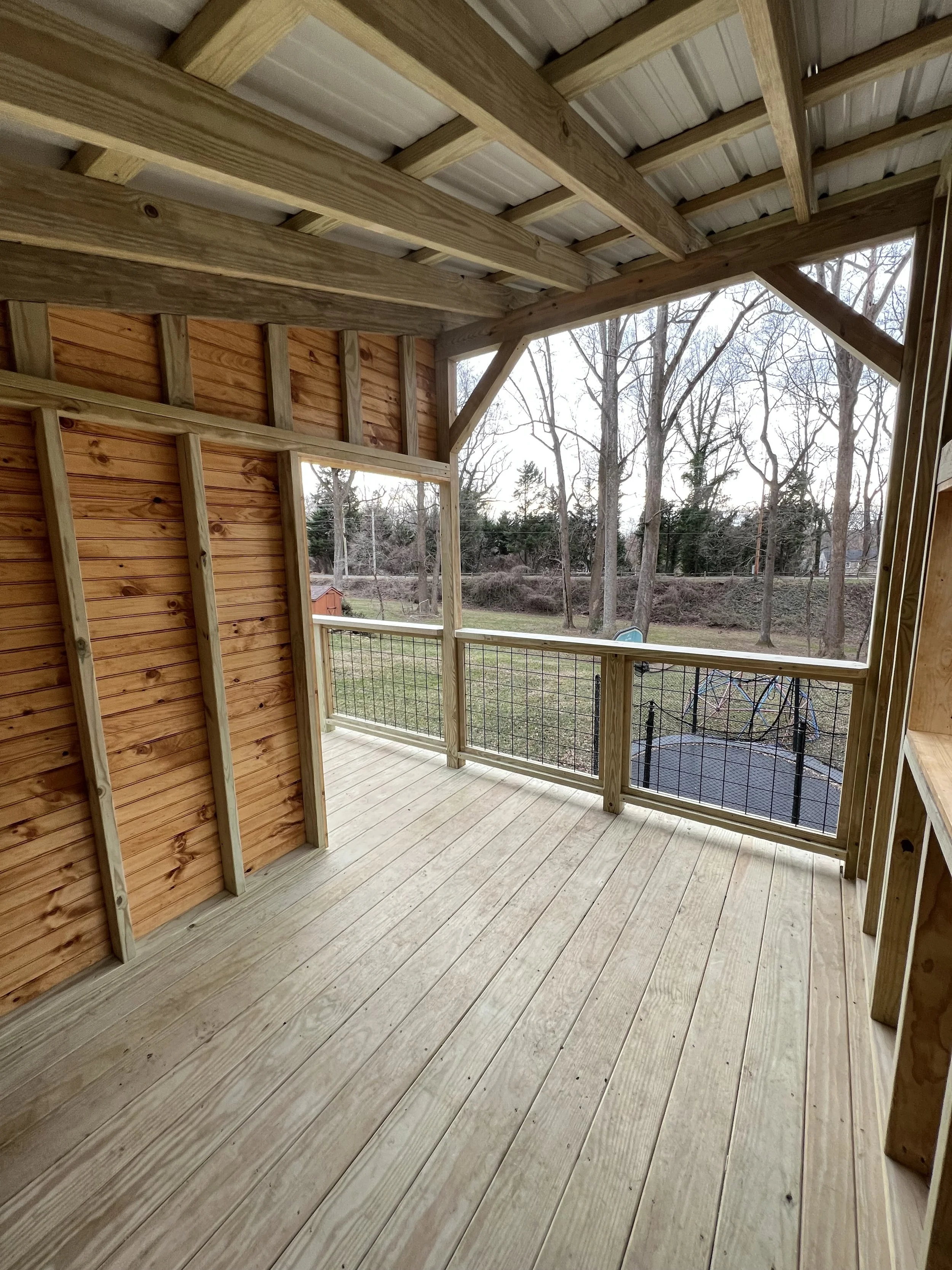 View of a newly constructed outdoor balcony with wooden flooring and railings, partial wooden wall, and a view of trees and backyard.