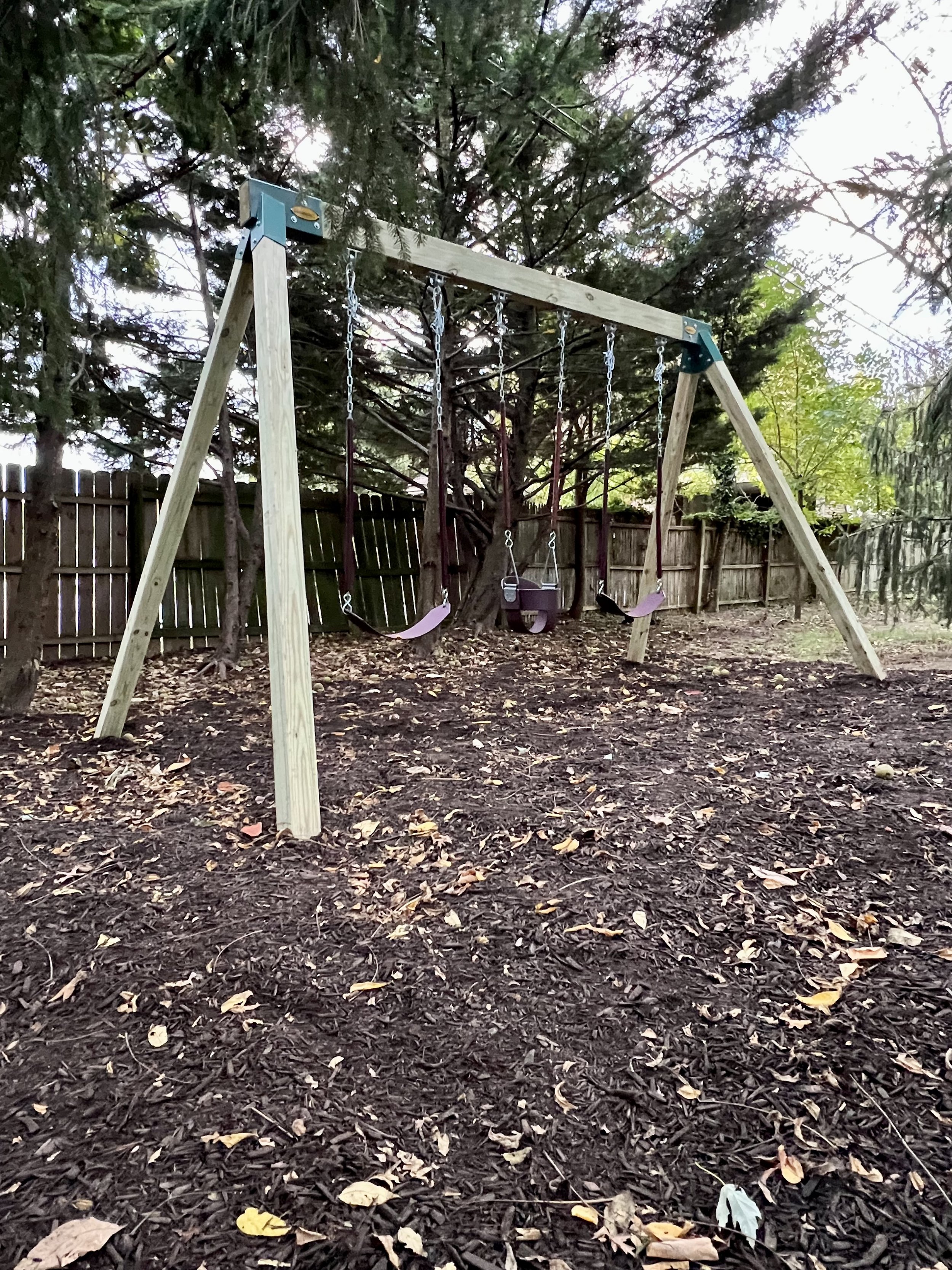 A wooden swing set with three swings, situated outdoors on a dirt ground covered with fallen leaves. Fenced trees are visible in the background.