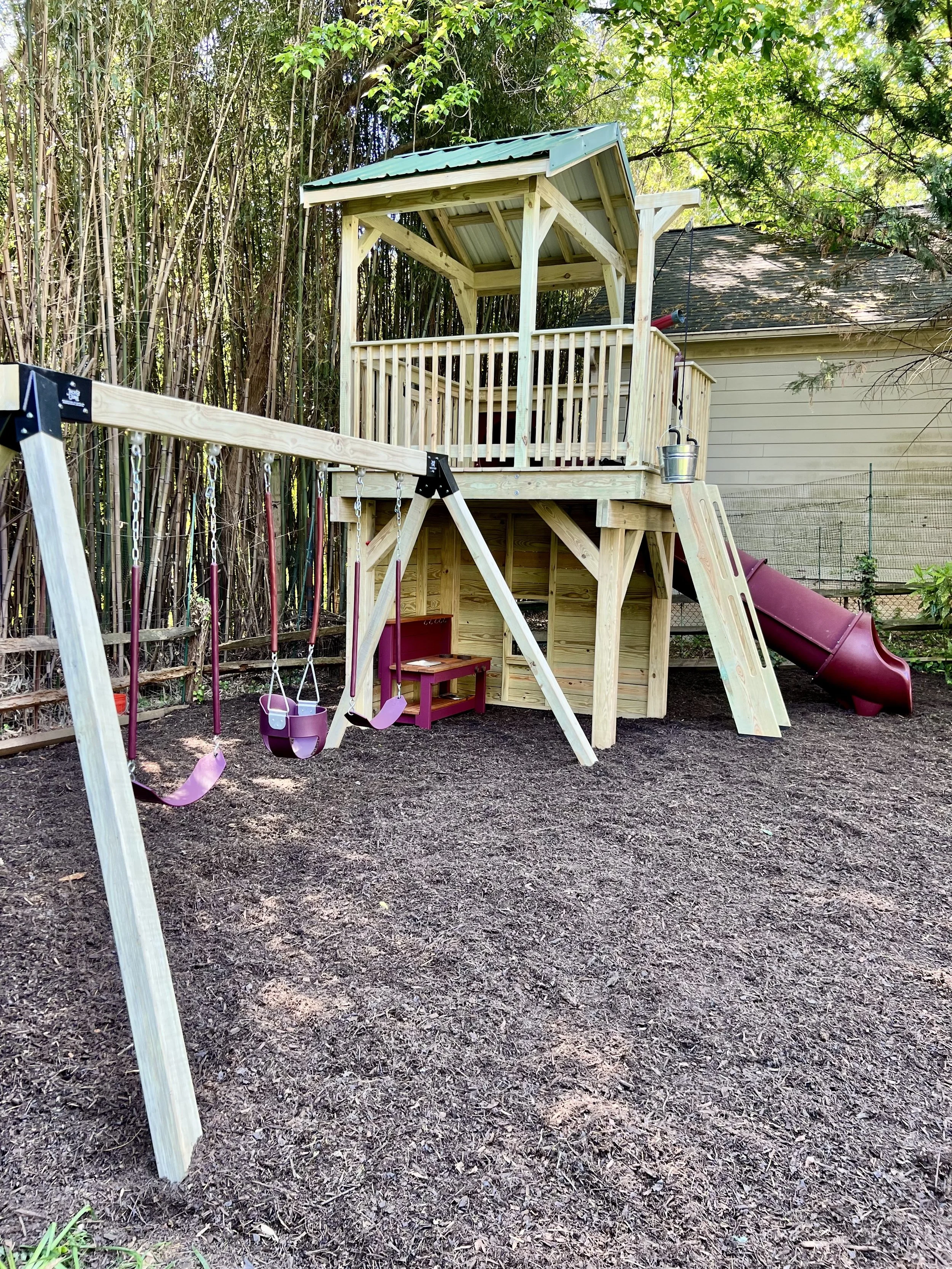 A backyard playground with a wooden treehouse, a swing set with pink seats, and a slide, set on a dirt ground with a fence and trees in the background.