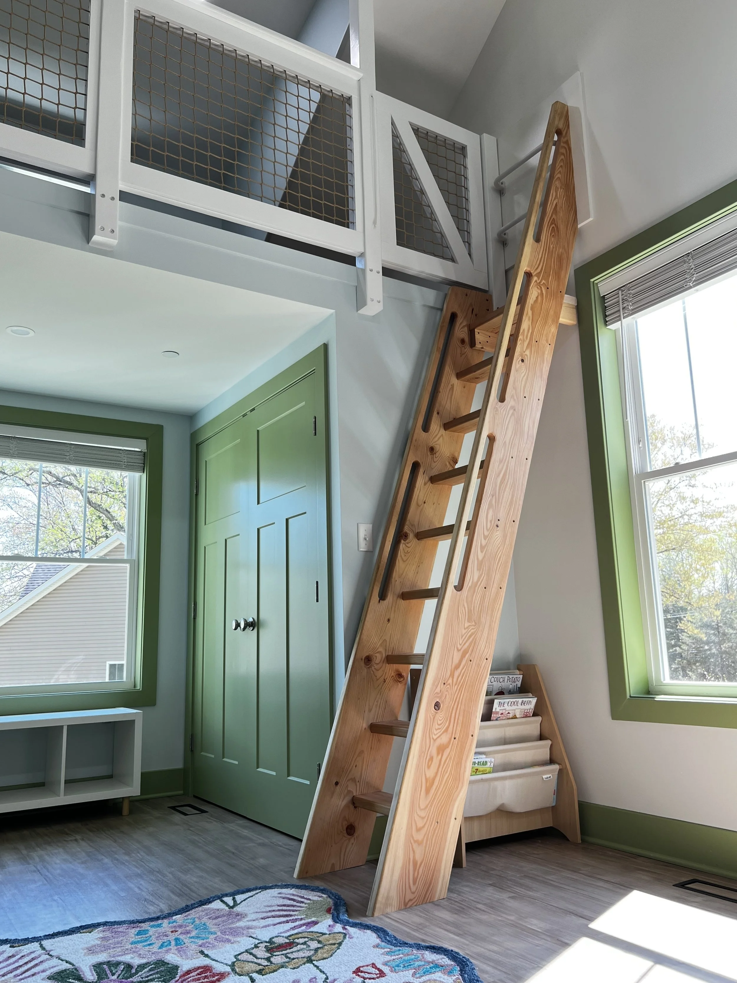 Interior of a room with a wooden loft ladder leading up to a small loft area, framed by green window trim and a matching door. The room has large windows with natural light and a colorful rug on the floor.