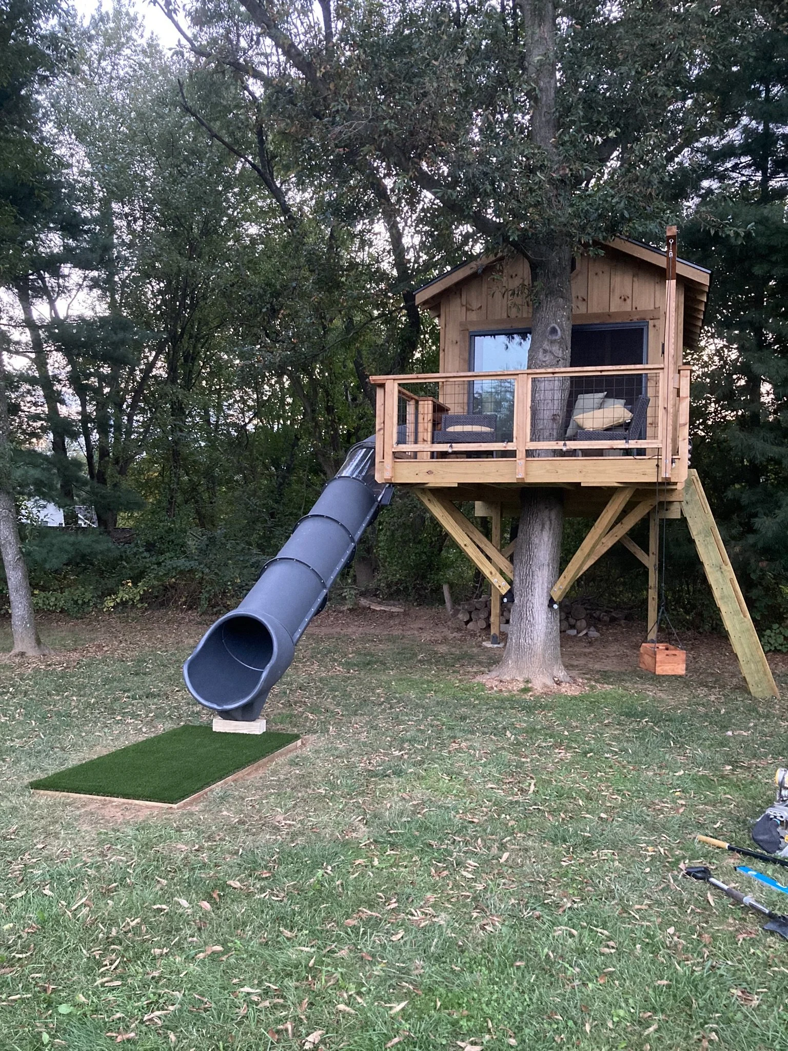A treehouse built around a large tree in a backyard, with a wooden deck, glass door, and a black sliding screen. A black slide curves down from the deck, and a swing hangs from the tree. The netting on the deck surrounds the tree, and there are outdoor chairs and a pillow on the deck. The yard has grass, scattered leaves, and trees in the background.