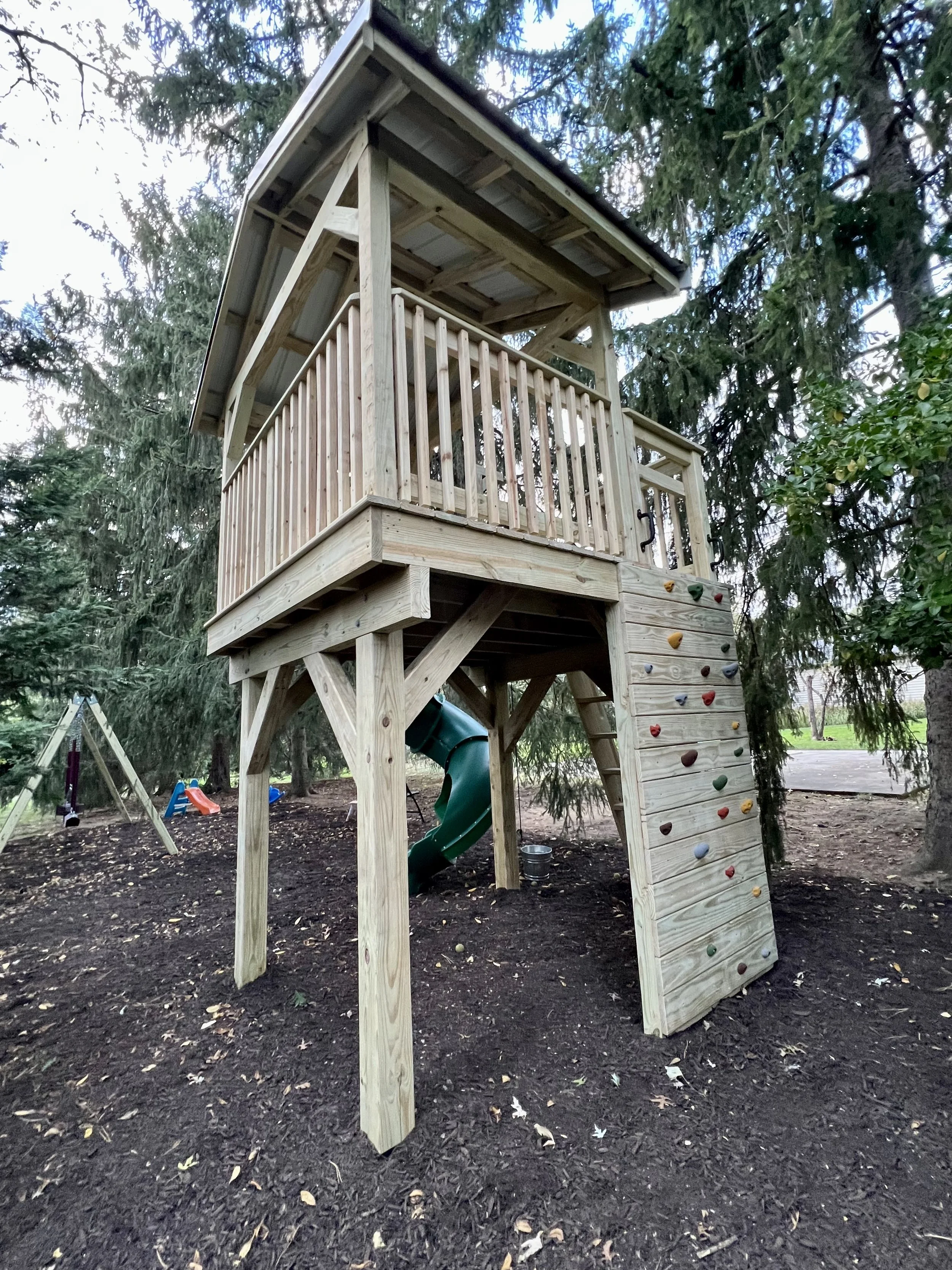 A wooden treehouse with a slide and climbing wall in a backyard, surrounded by tall trees.