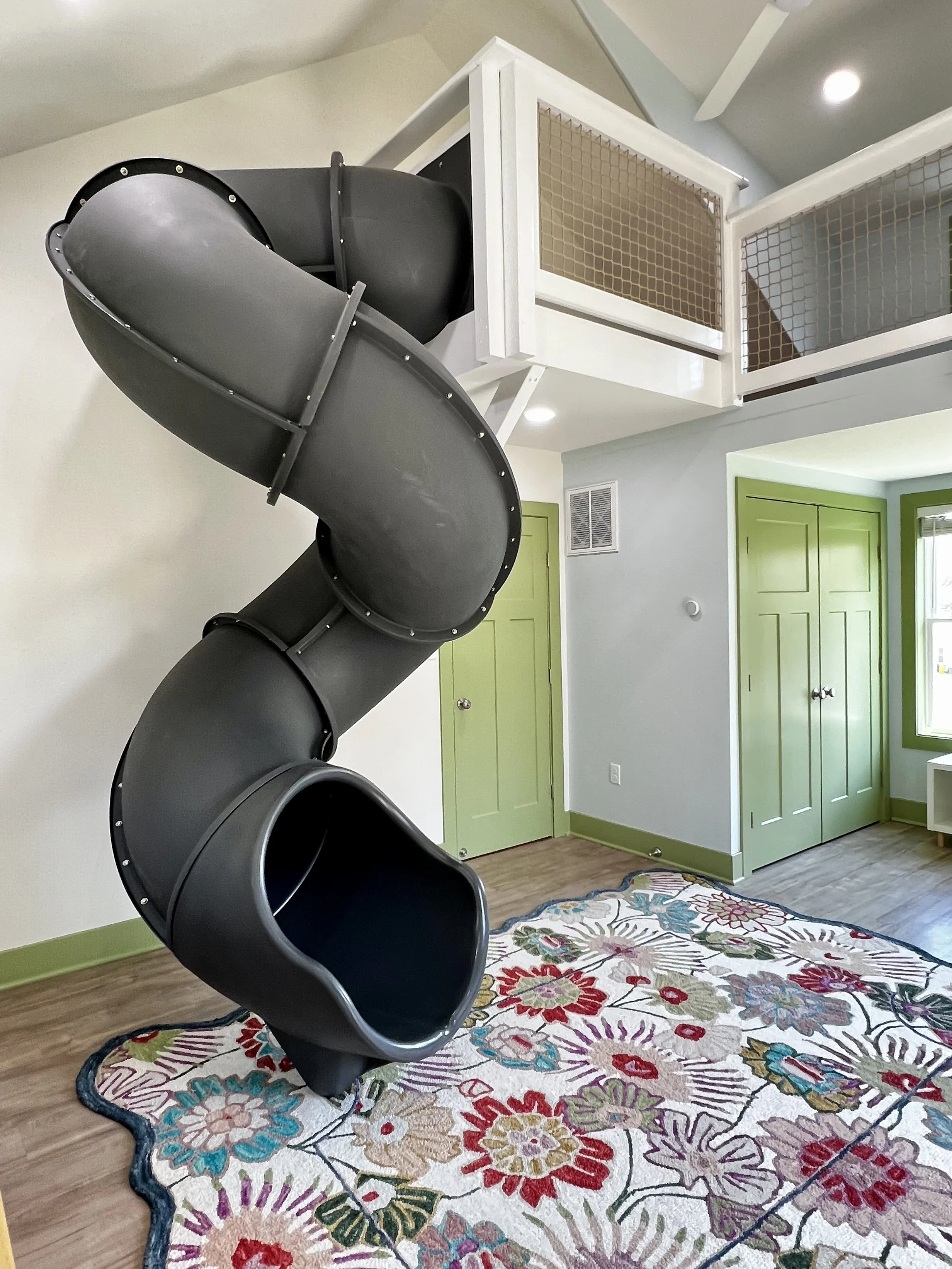 A black spiral slide in a brightly colored room with green doors, a floral area rug, and a ceiling with recessed lighting.