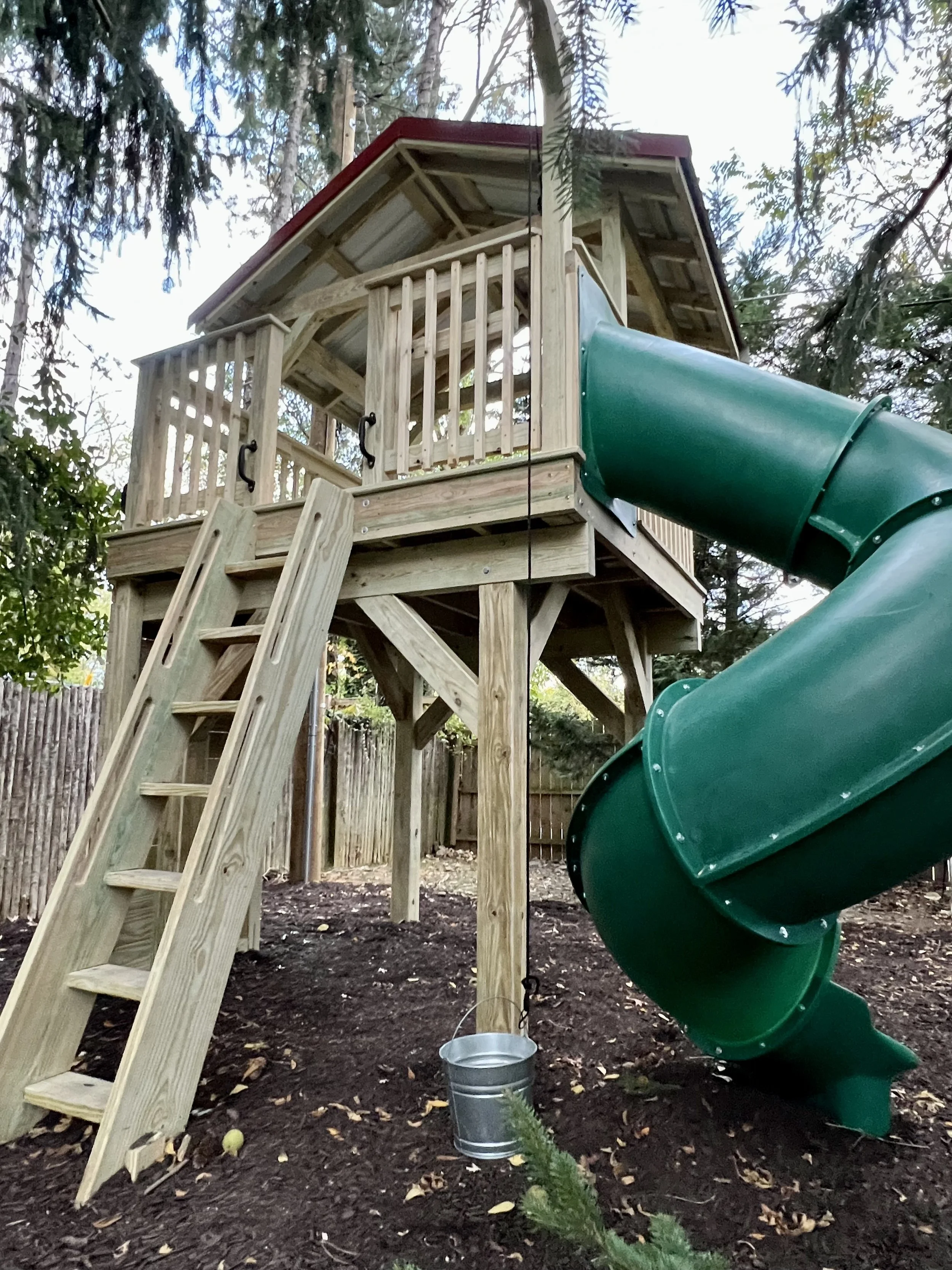 A wooden playhouse with stairs and a slide in a backyard, surrounded by trees and a wooden fence.
