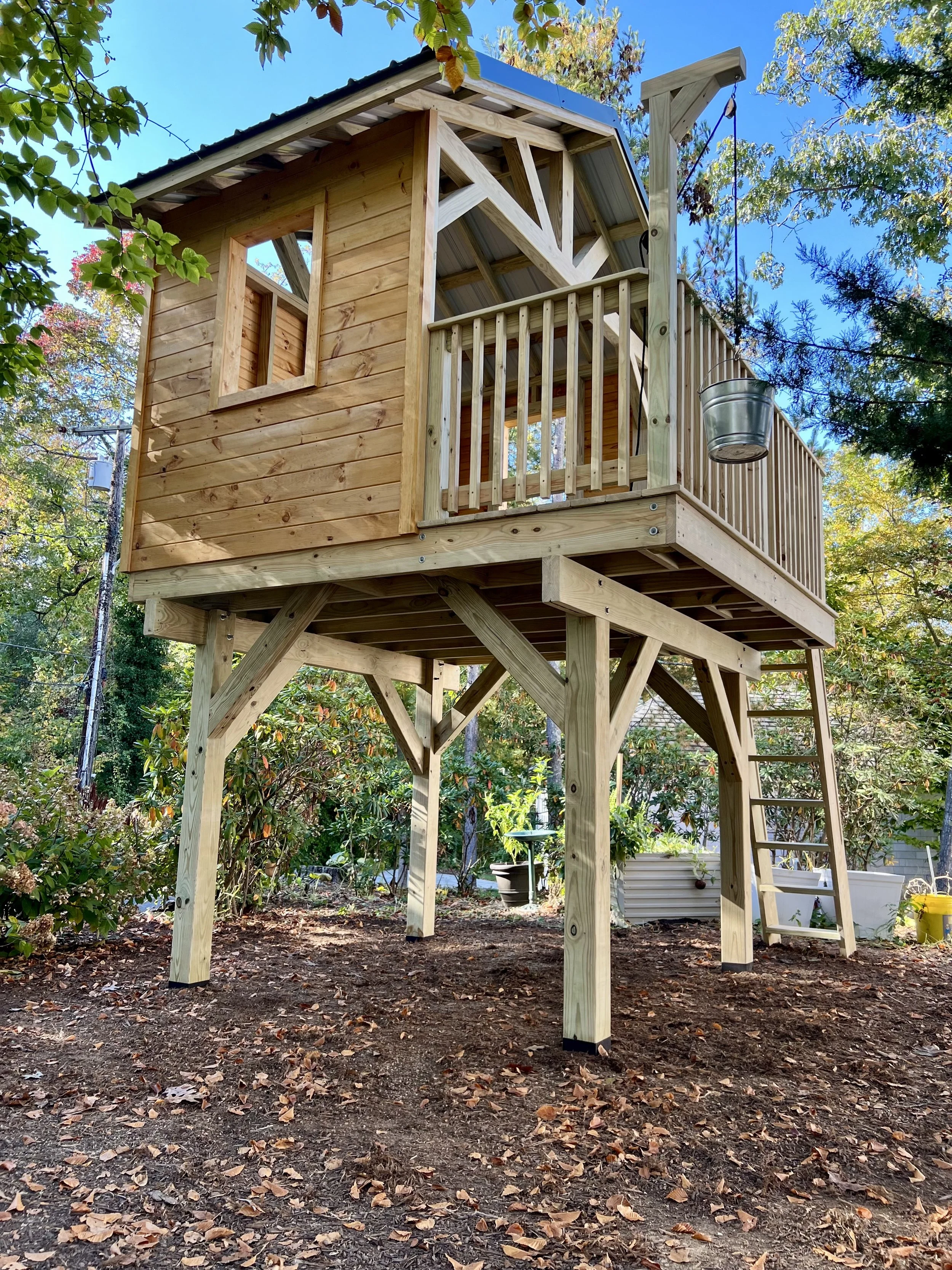 A wooden play structure, featuring a small balcony with a railing, an open window, and with a ladder leading up to it, situated in a backyard with trees and fallen leaves.