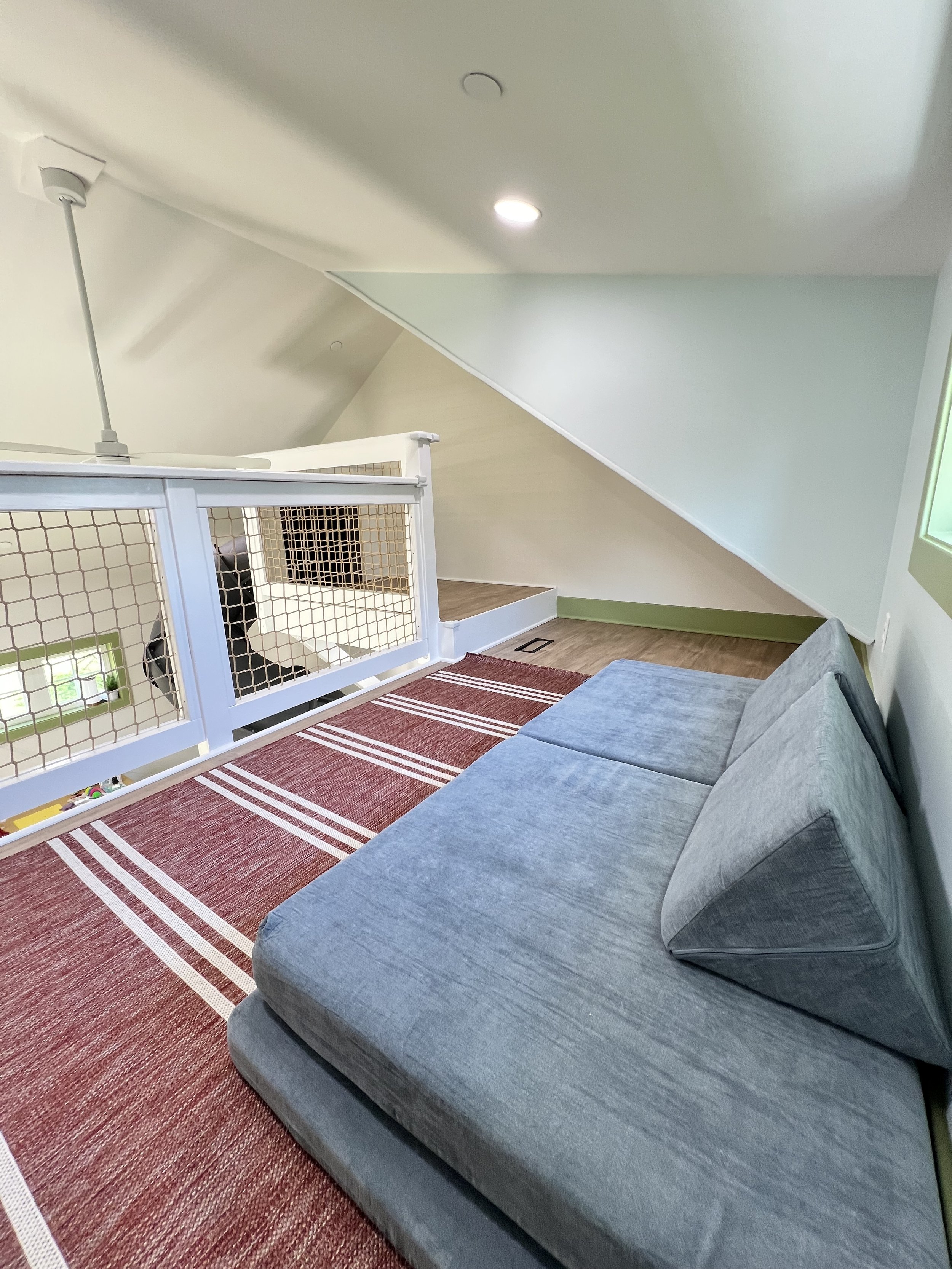 Living room with a gray sofa, a red striped rug, and a white railing with wooden lattice, overlooking a staircase. The ceiling has recessed lighting and a ceiling fan.