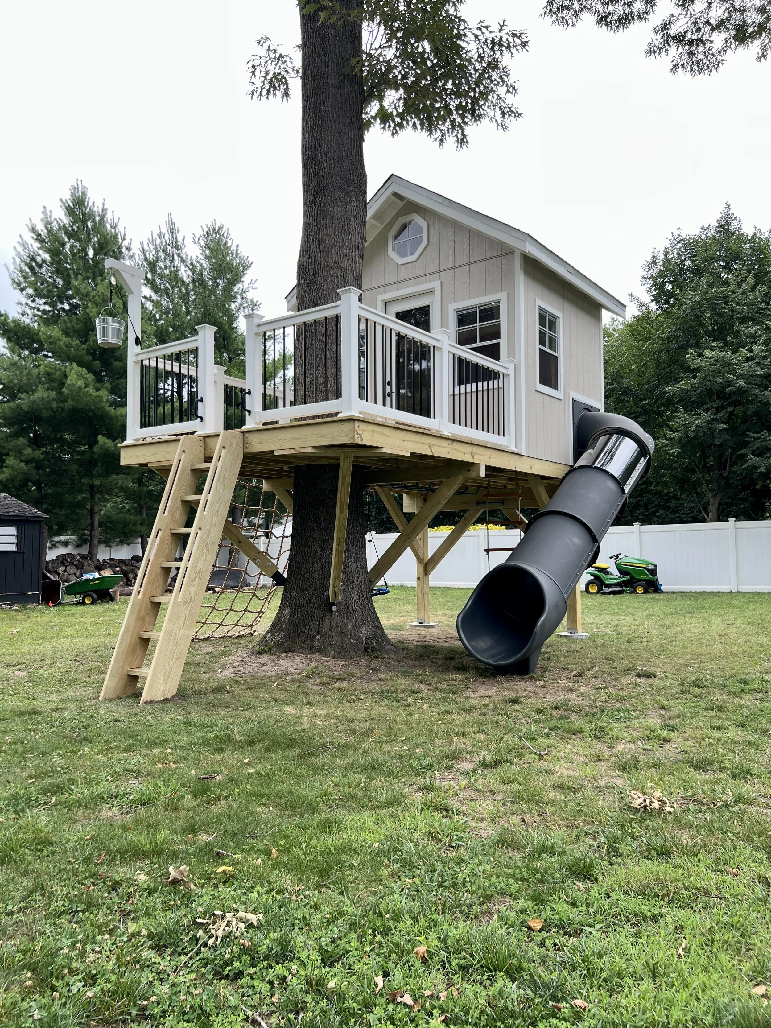 A treehouse built around a large tree in a backyard with a slide, rope ladder, and railing, with a toy tractor and a small black shed in the yard.