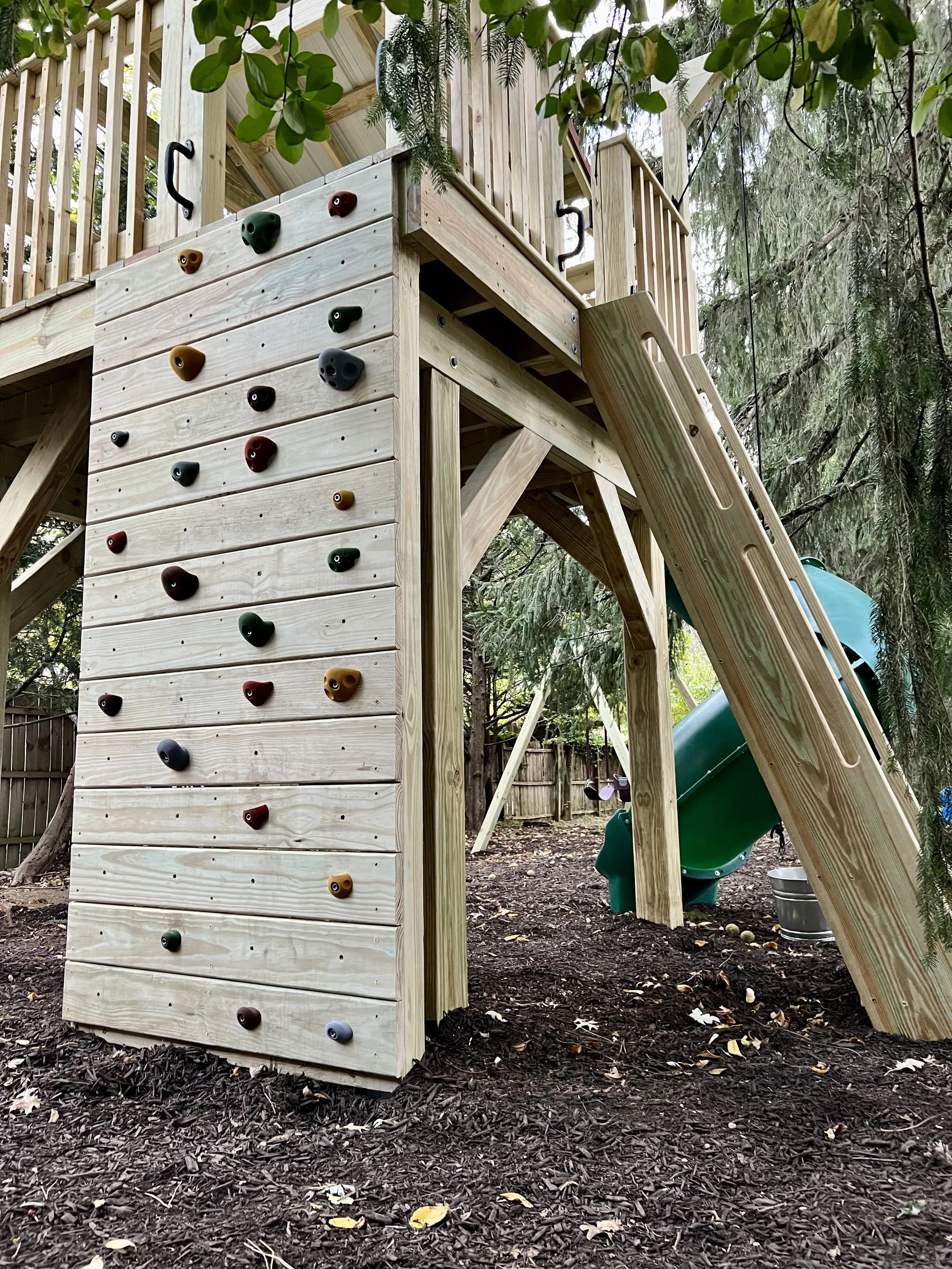 A wooden outdoor play structure with a climbing wall and a slide, situated in a wooded backyard.