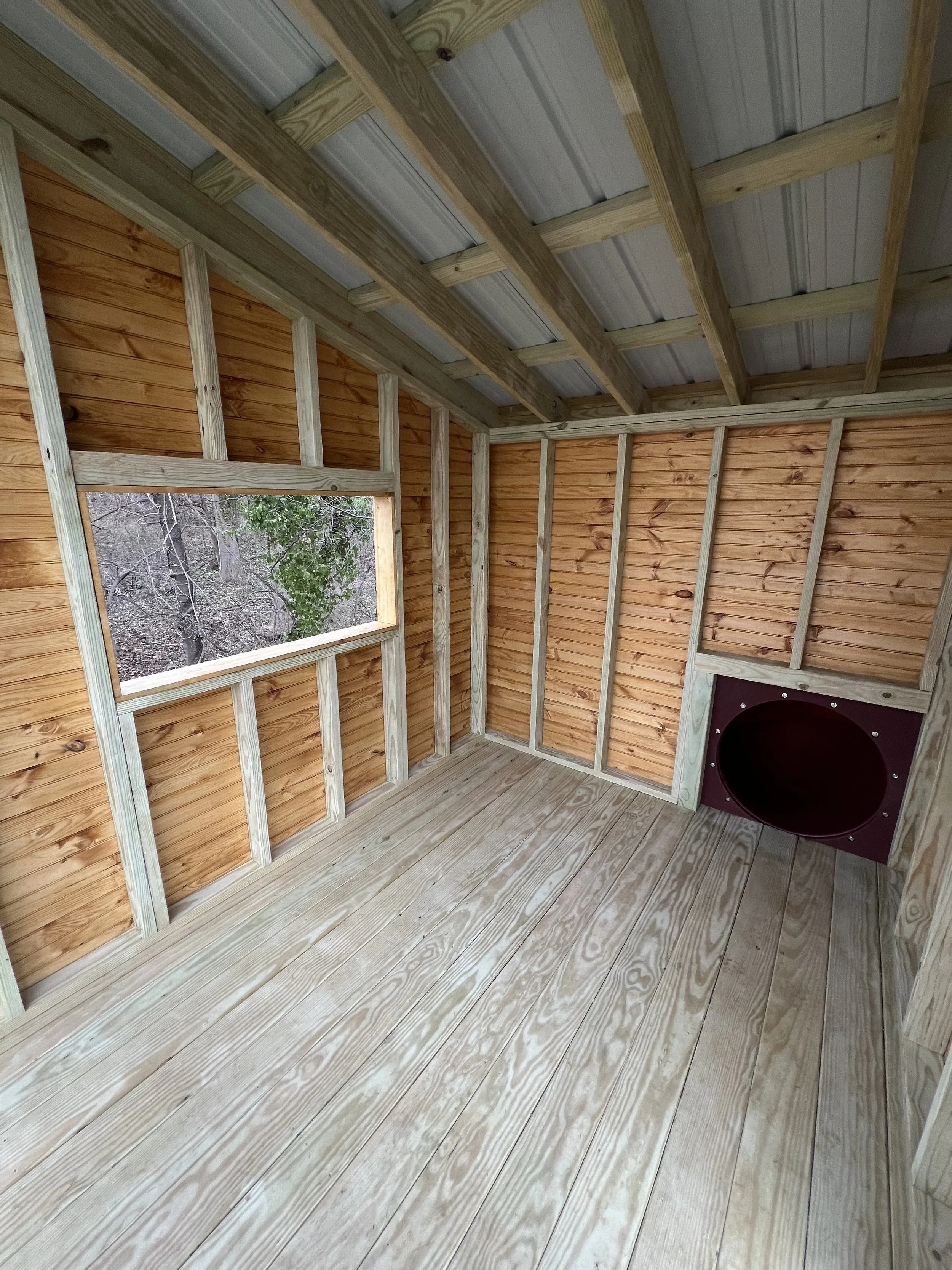 Interior of a partially built wooden shed with exposed framing, a window, and an oval opening for a wood stove or heater, with wood walls, ceiling, and floor.