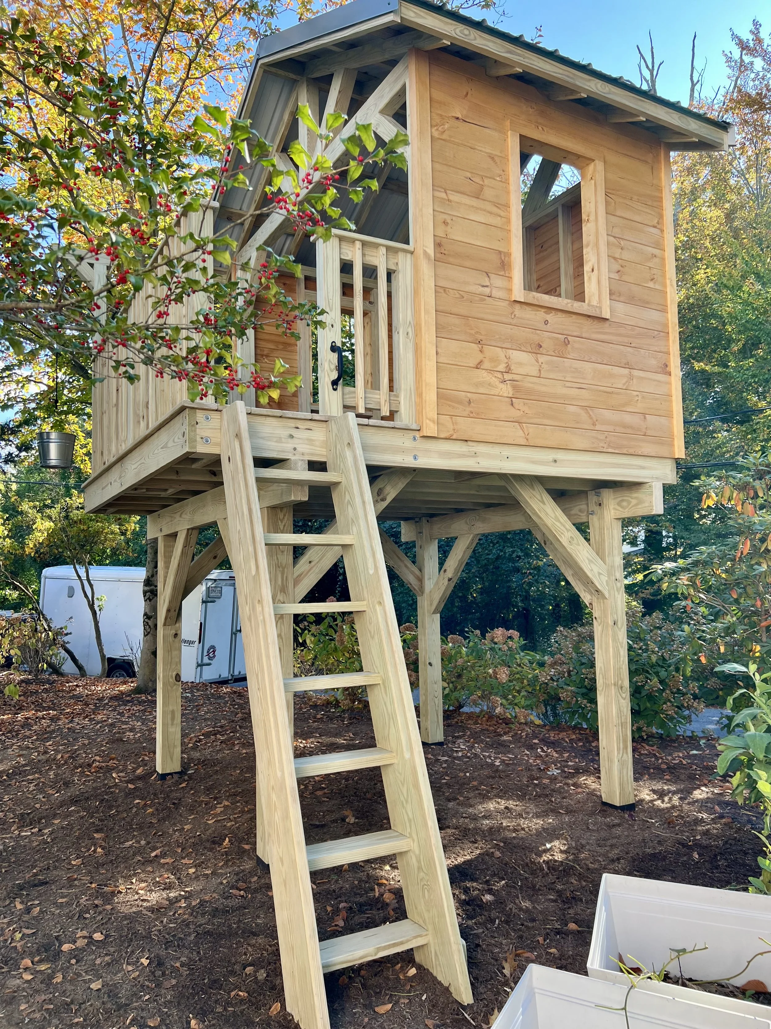 A small wooden play structure, with a ladder leading up to a deck and an unfinished door and window frame. The scene is set outdoors in a garden with trees and fallen leaves.