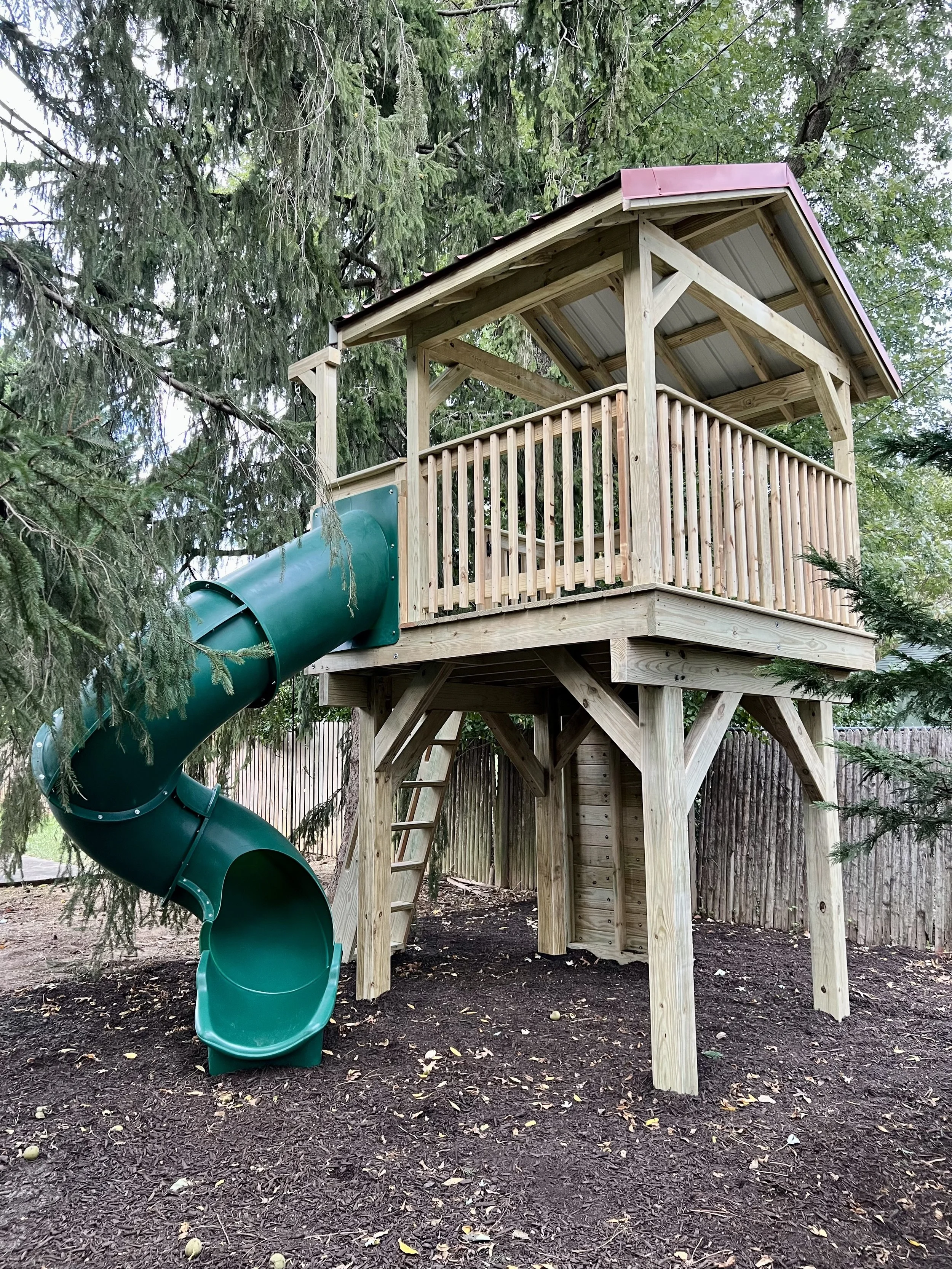 Wooden playground structure with a green tube slide surrounded by trees and fenced yard, on mulch ground.