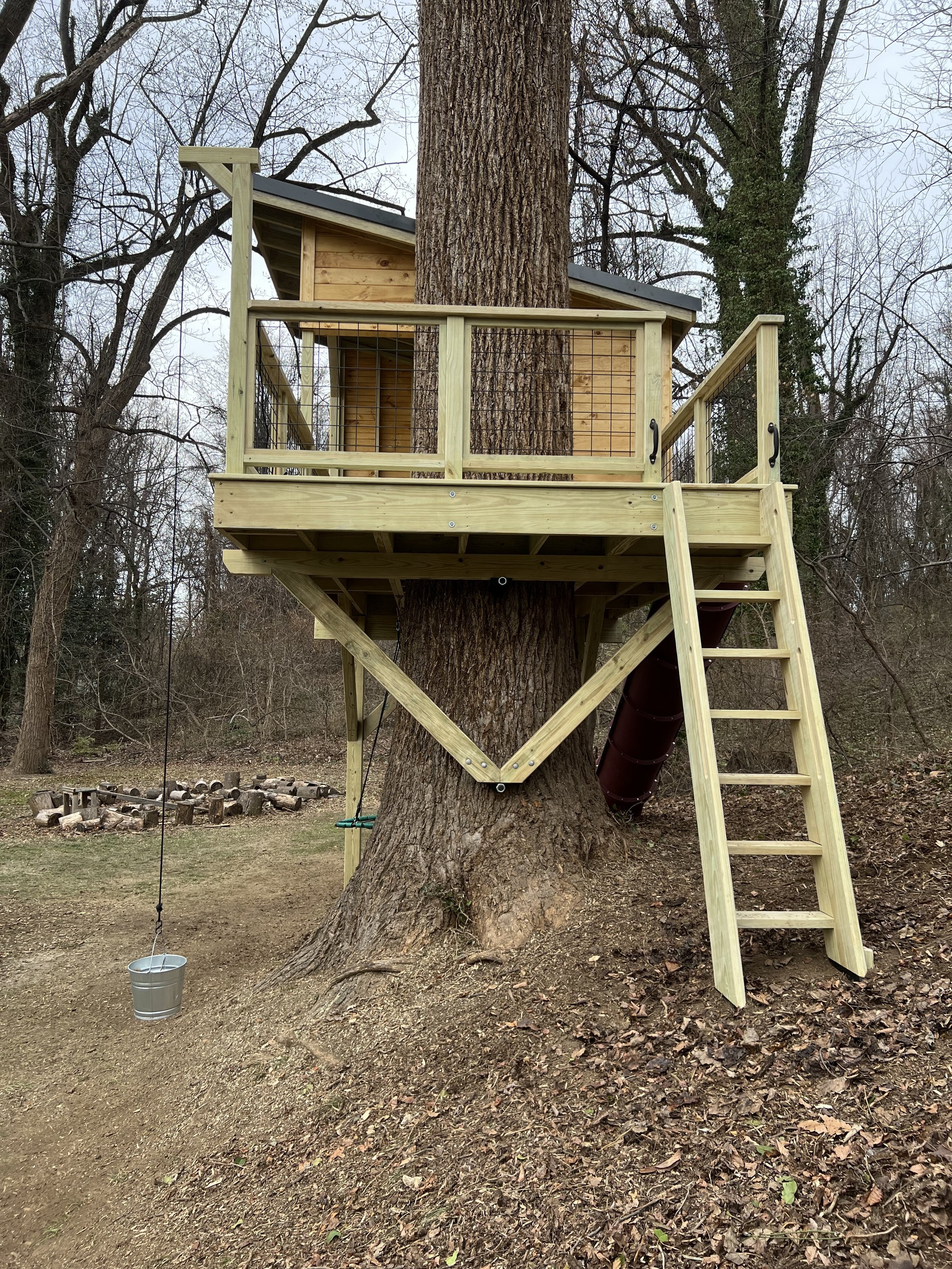 A treehouse built around a large tree, with a wooden deck and ladder, in a wooded area with leafless trees, and a swing hanging from a rope on the left side.