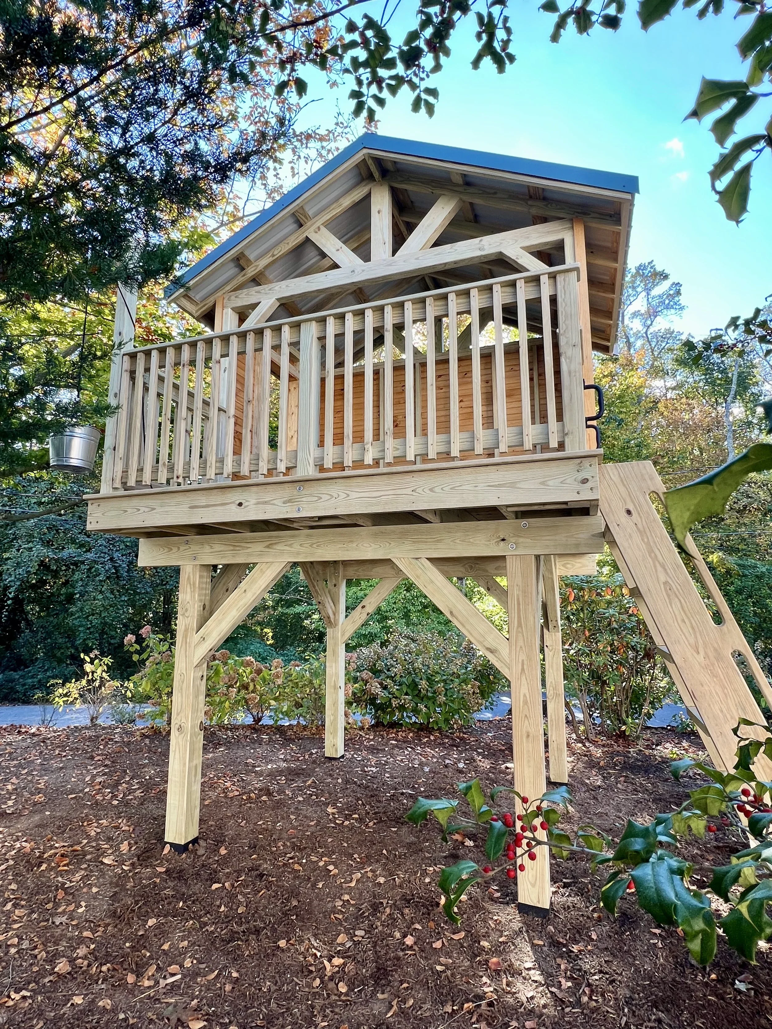 A play structure constructed with wooden planks, featuring a balcony with a railing and a sloped roof, elevated above the ground in a wooded area with trees and bushes surrounding it, and a slide attached to its side.