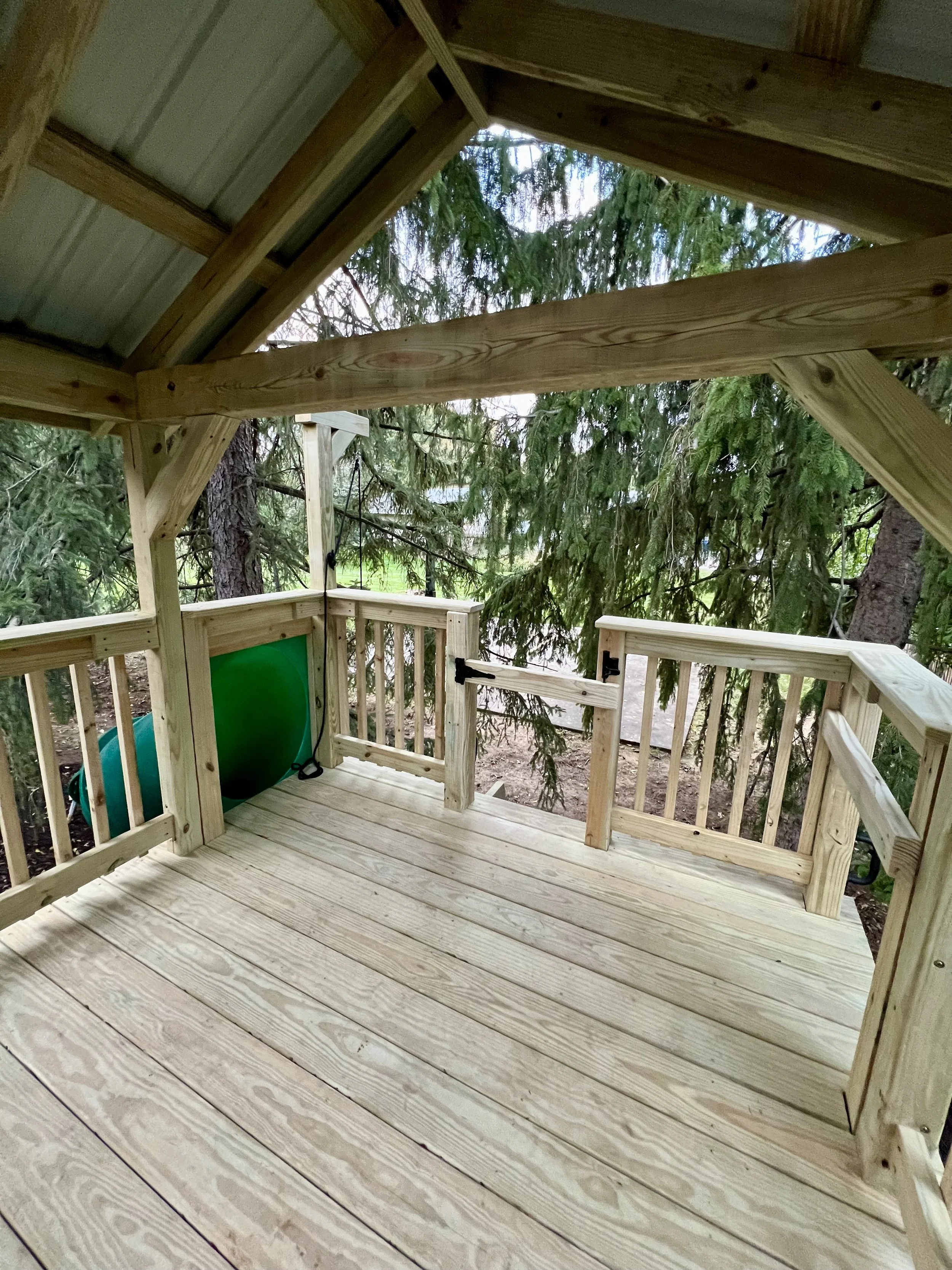 A wooden outdoor deck with a railing, surrounded by trees, and part of a tree house or similar structure under construction.