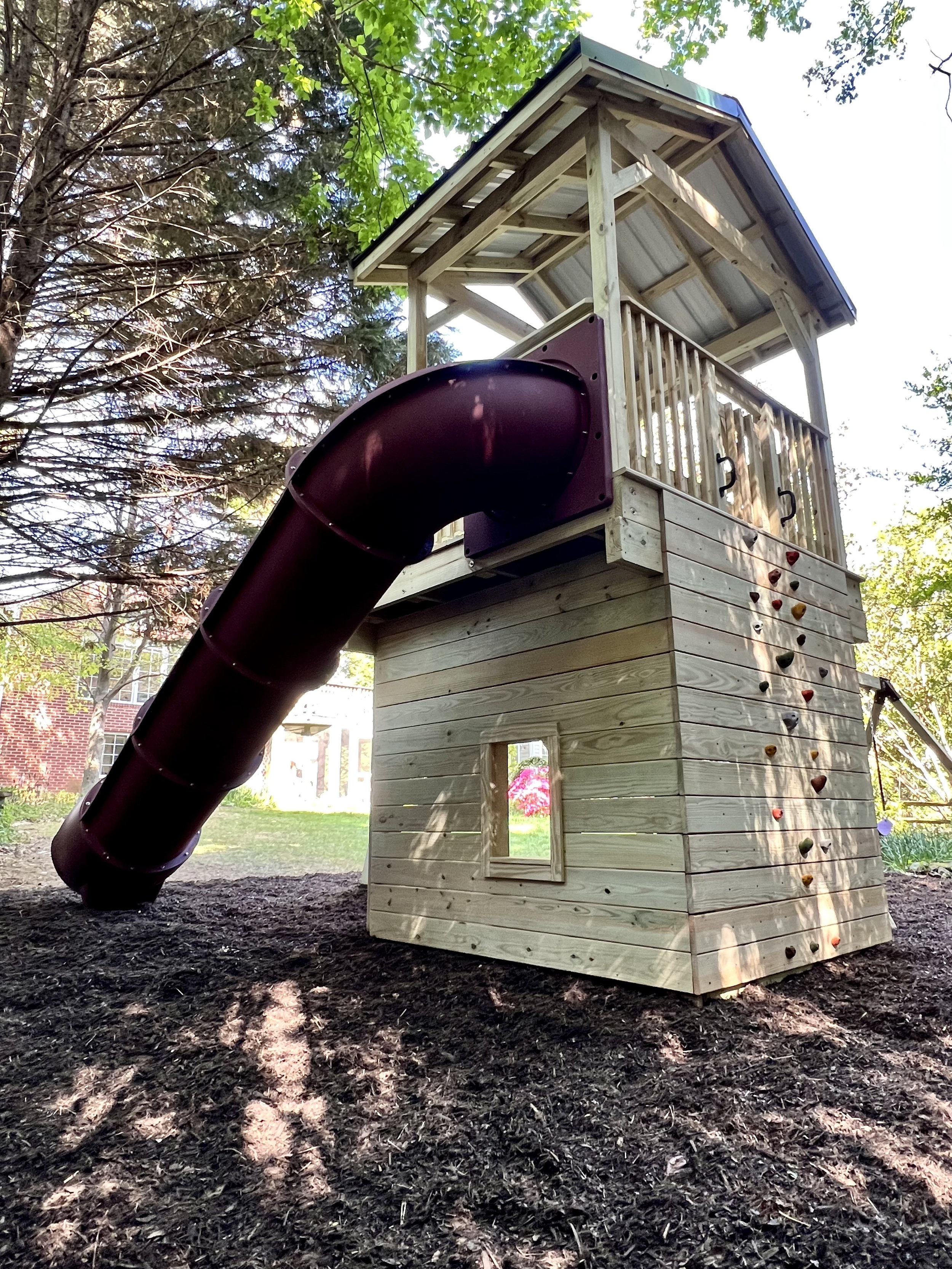 Wooden play structure with a slide and climbing wall in a backyard with trees and grass.