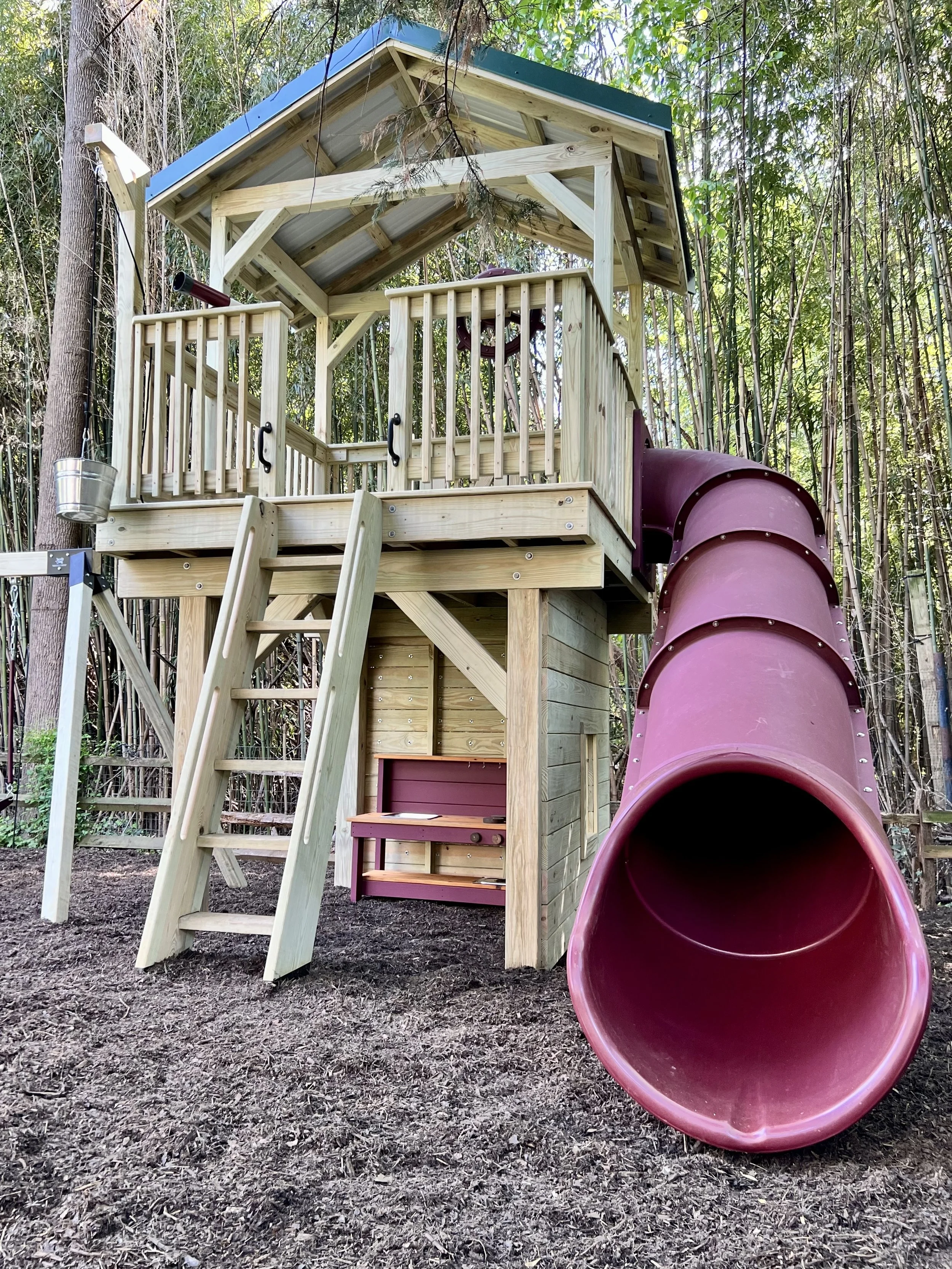 A wooden play structure with a slide and a small porch, set in a wooded area with trees and bushes in the background.