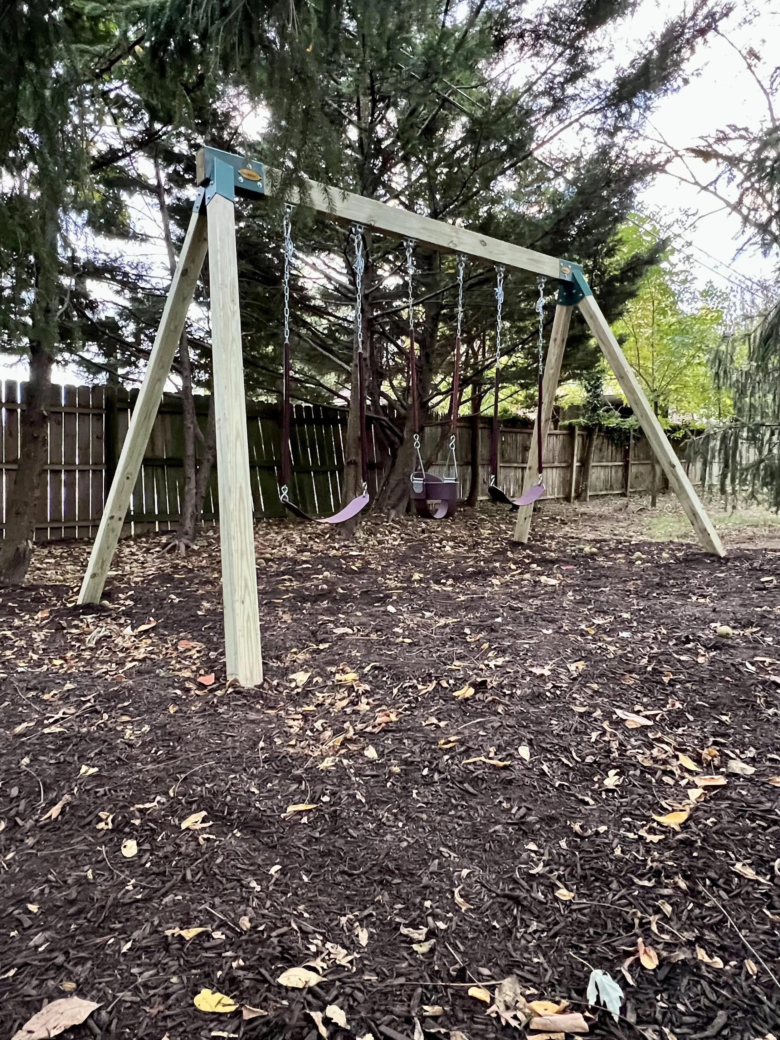 A wooden swing set with two swings and a toddler swing in a backyard, surrounded by trees and fallen leaves.