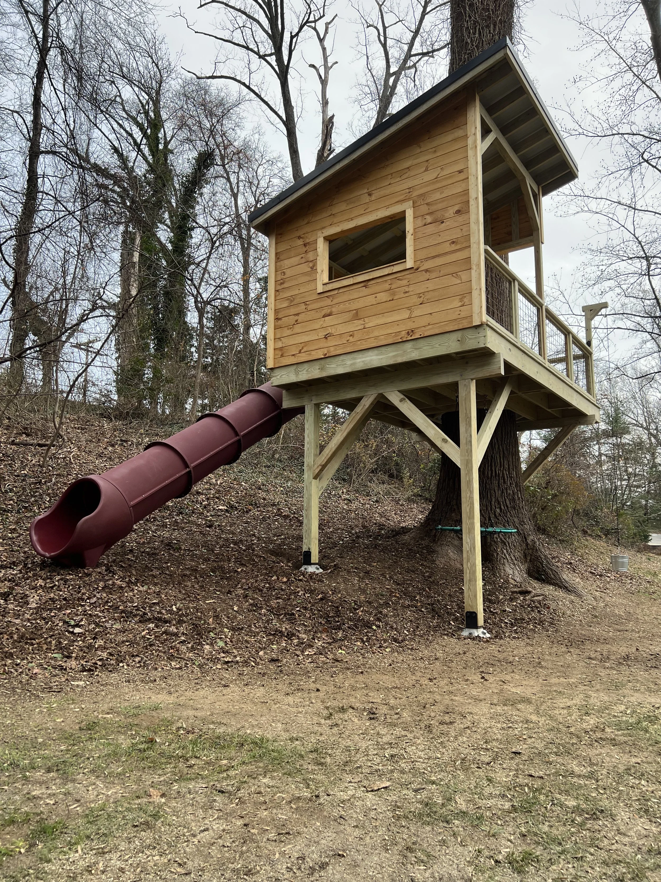 The image shows a treehouse built around a large tree in a wooded area. The treehouse has wooden siding, a sloped metal roof, and a wrap-around porch with a railing. There is a plastic tube slide connected to the treehouse, descending to the ground below.
