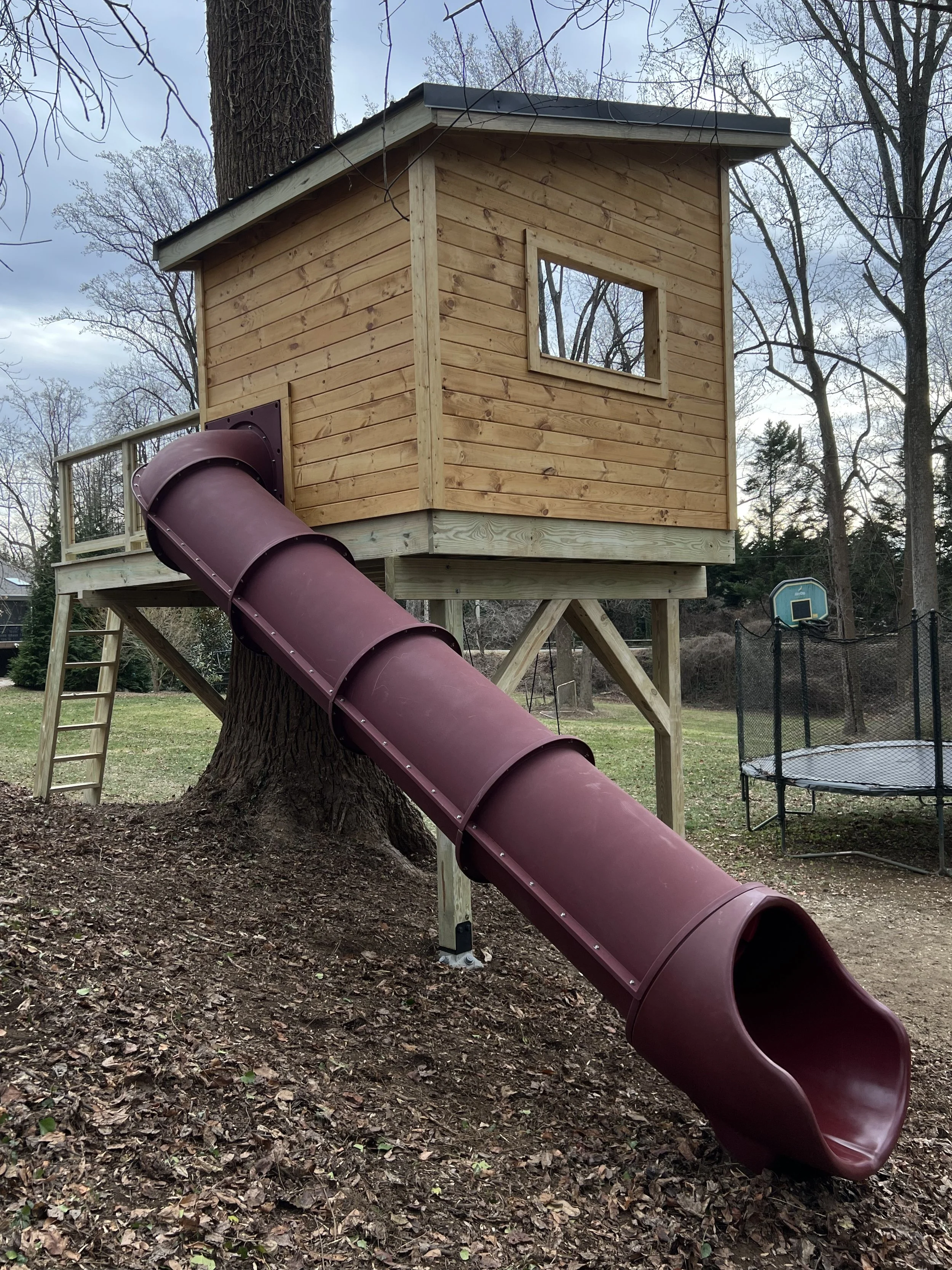 A wooden play structure with a window, a chimney, and a spiral slide attached, situated in a yard with trees and a trampoline nearby.