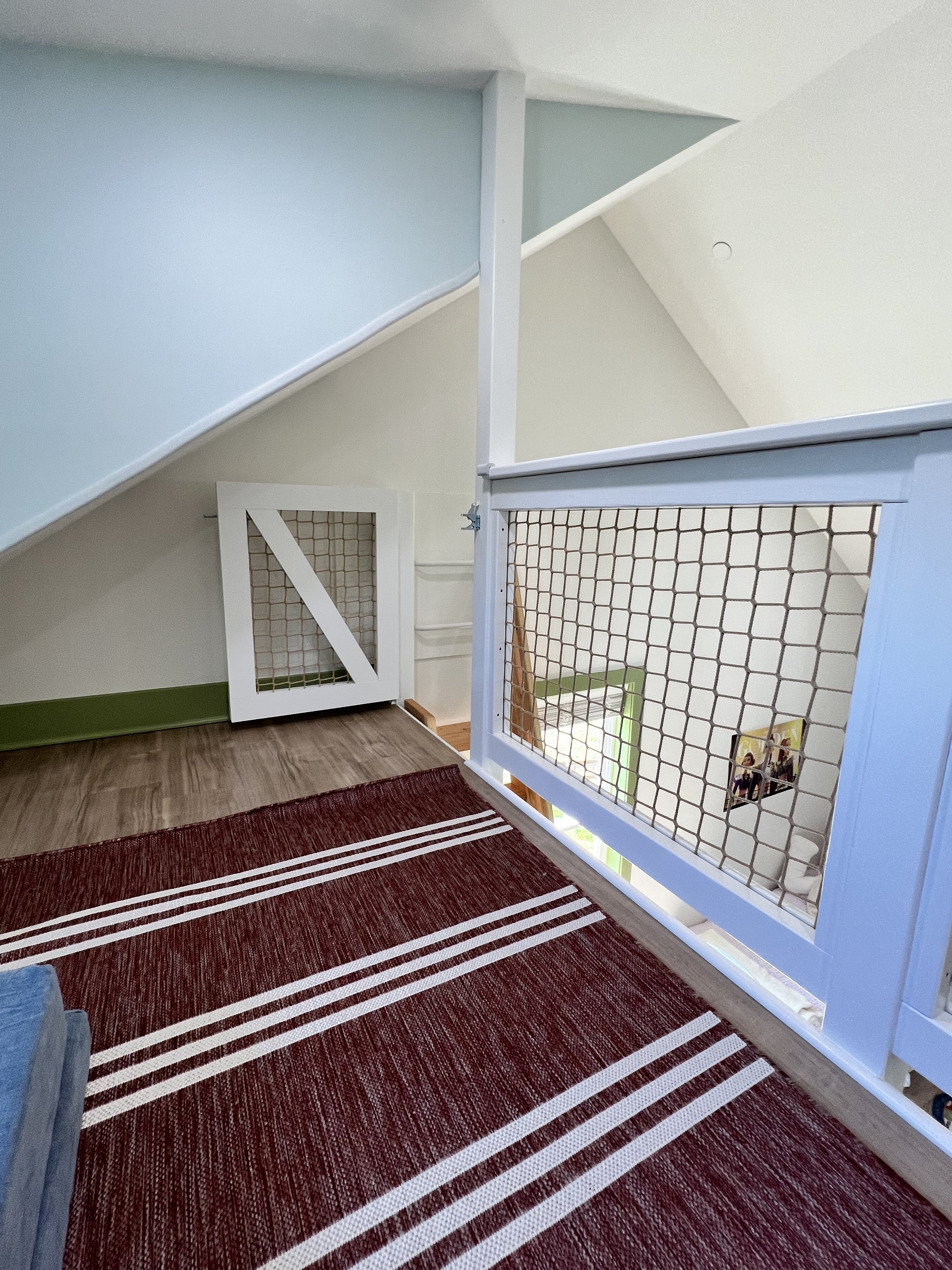 Interior view of a staircase area with a red striped rug on the wooden floor, white and green walls, a white wooden railing with a netted panel, and a small white door with a grid pattern.