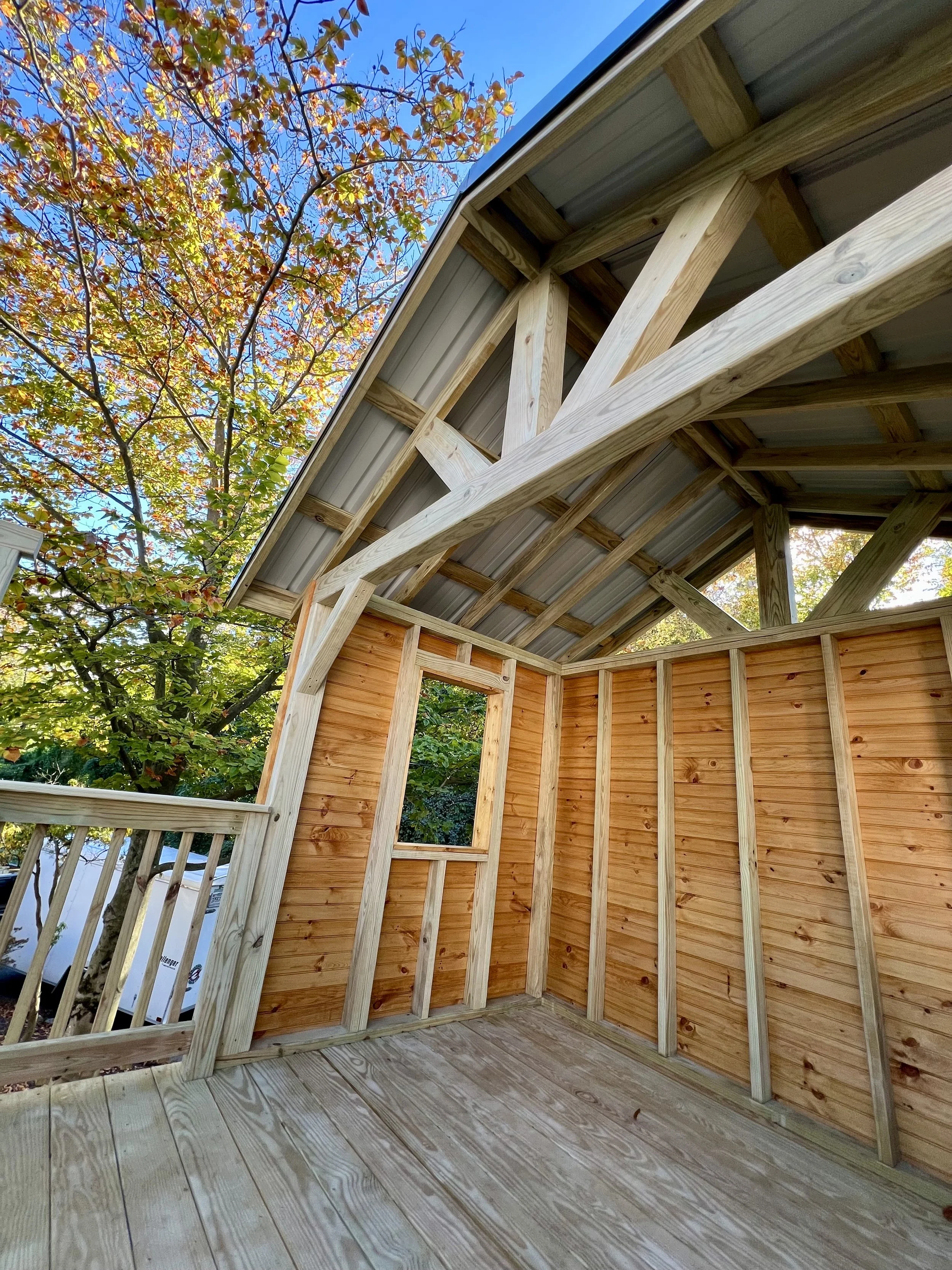 The image shows the interior of a wooden play structure, with a view of the roof supported by wooden beams and a partial wall with horizontal wooden siding. Outside, there are trees with autumn-colored leaves, and the sky is clear.