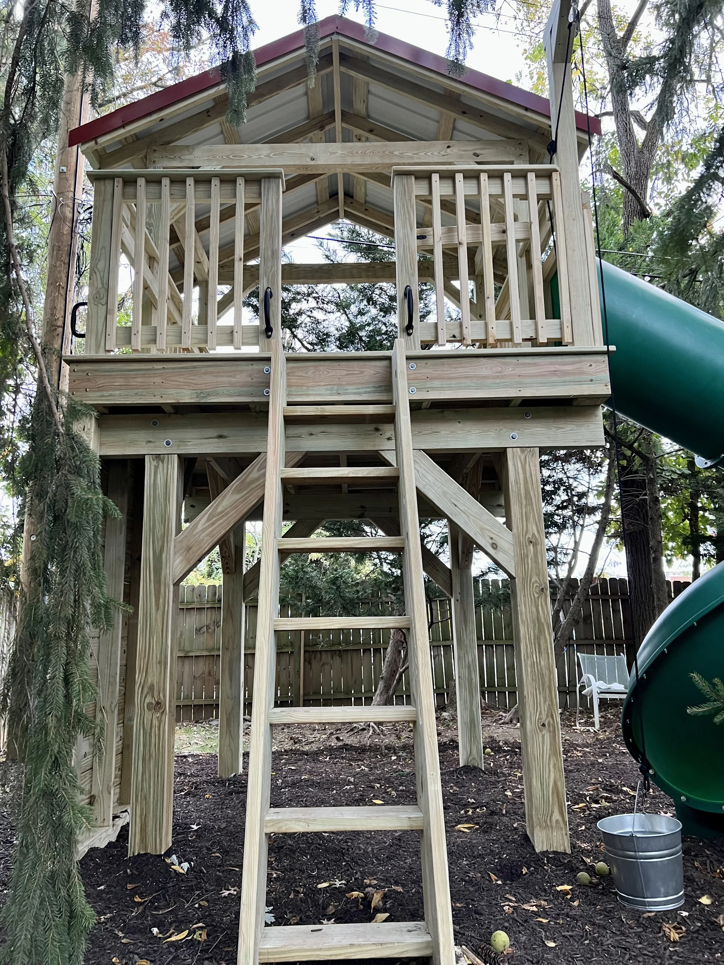 A wooden treehouse with a slide on the right side, built among trees, with a ladder leading up to it and surrounded by a fence.