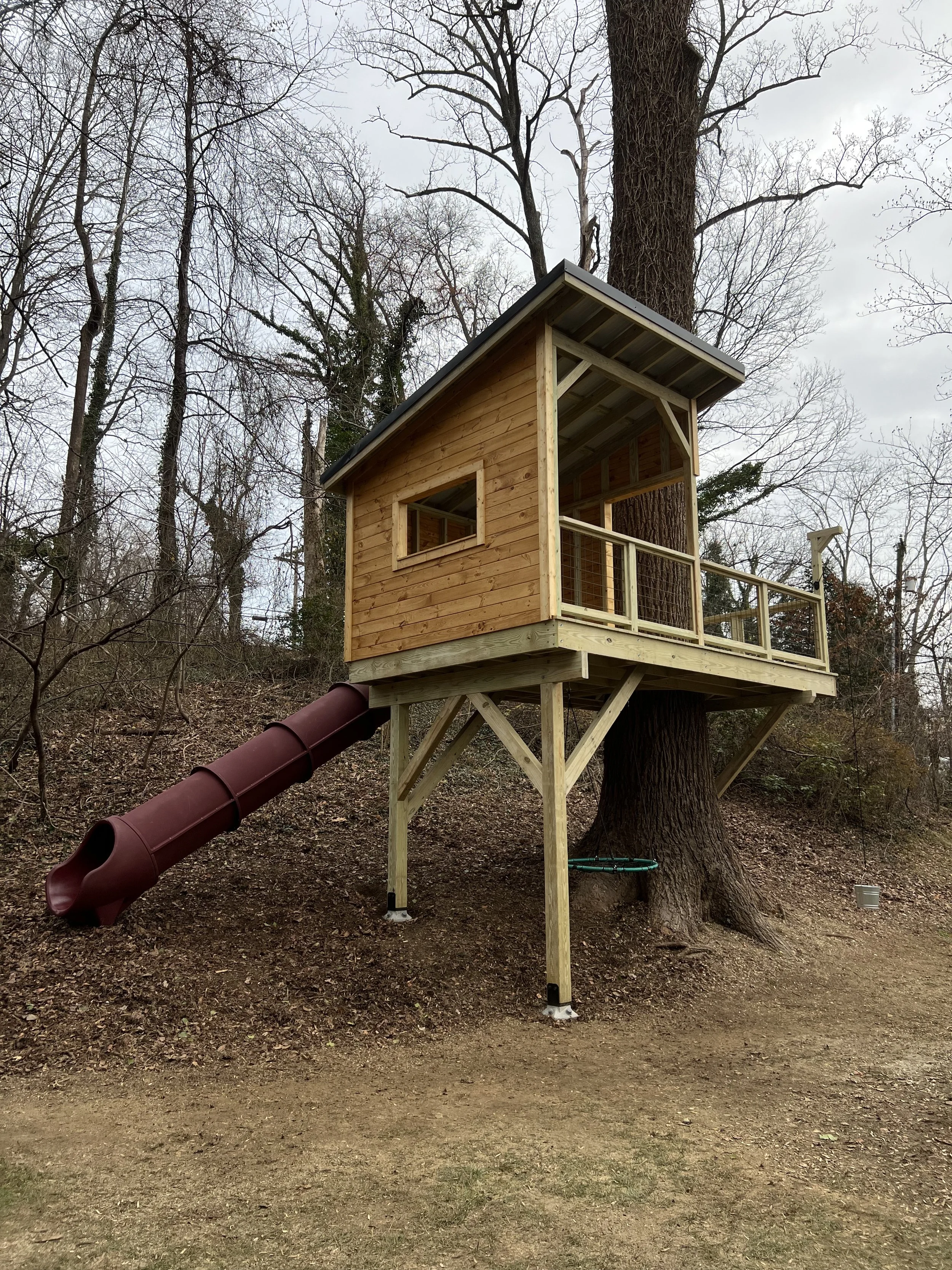 A treehouse in a wooded area, with a ladder, rope bucket, railing, and a slide attached to the back.