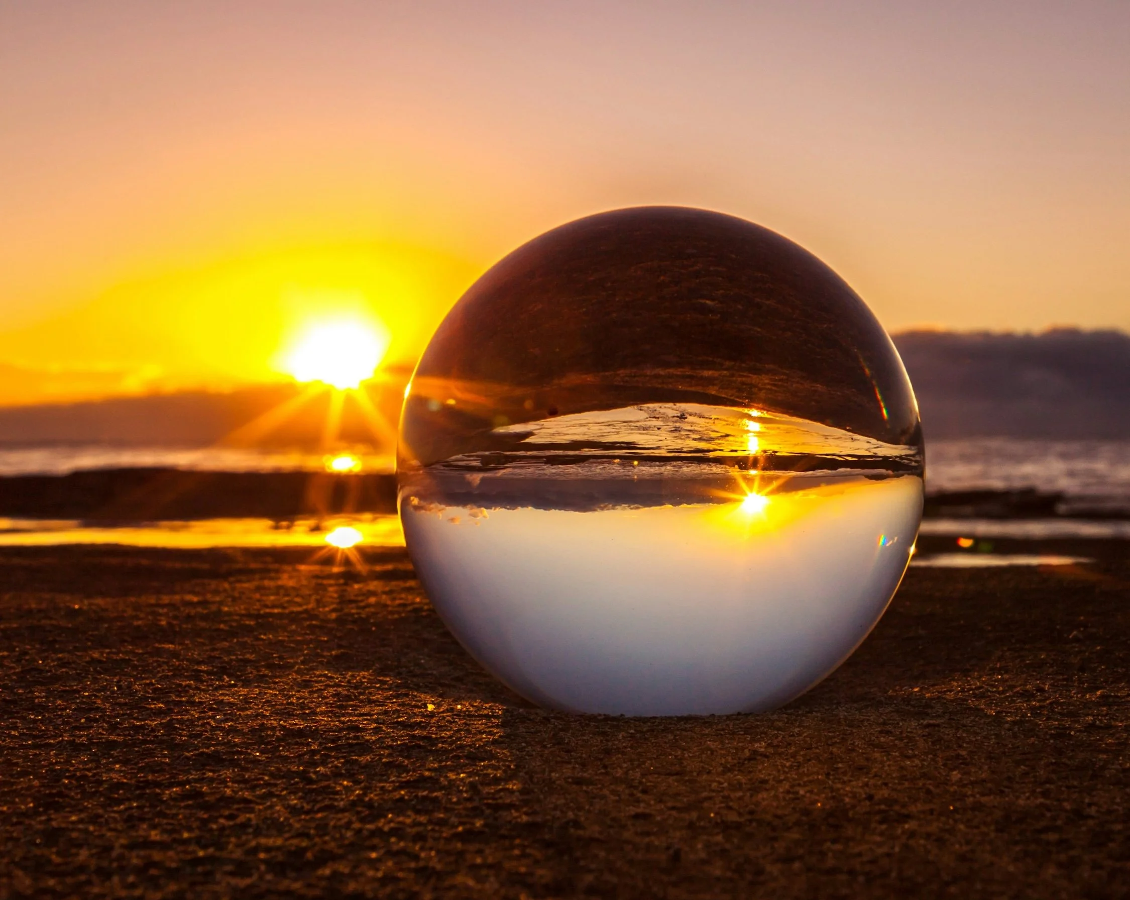 A beach at sunrise is shown blurred in the background. In the foreground is a glass ball sitting on the sand, reflecting the scene upside down.