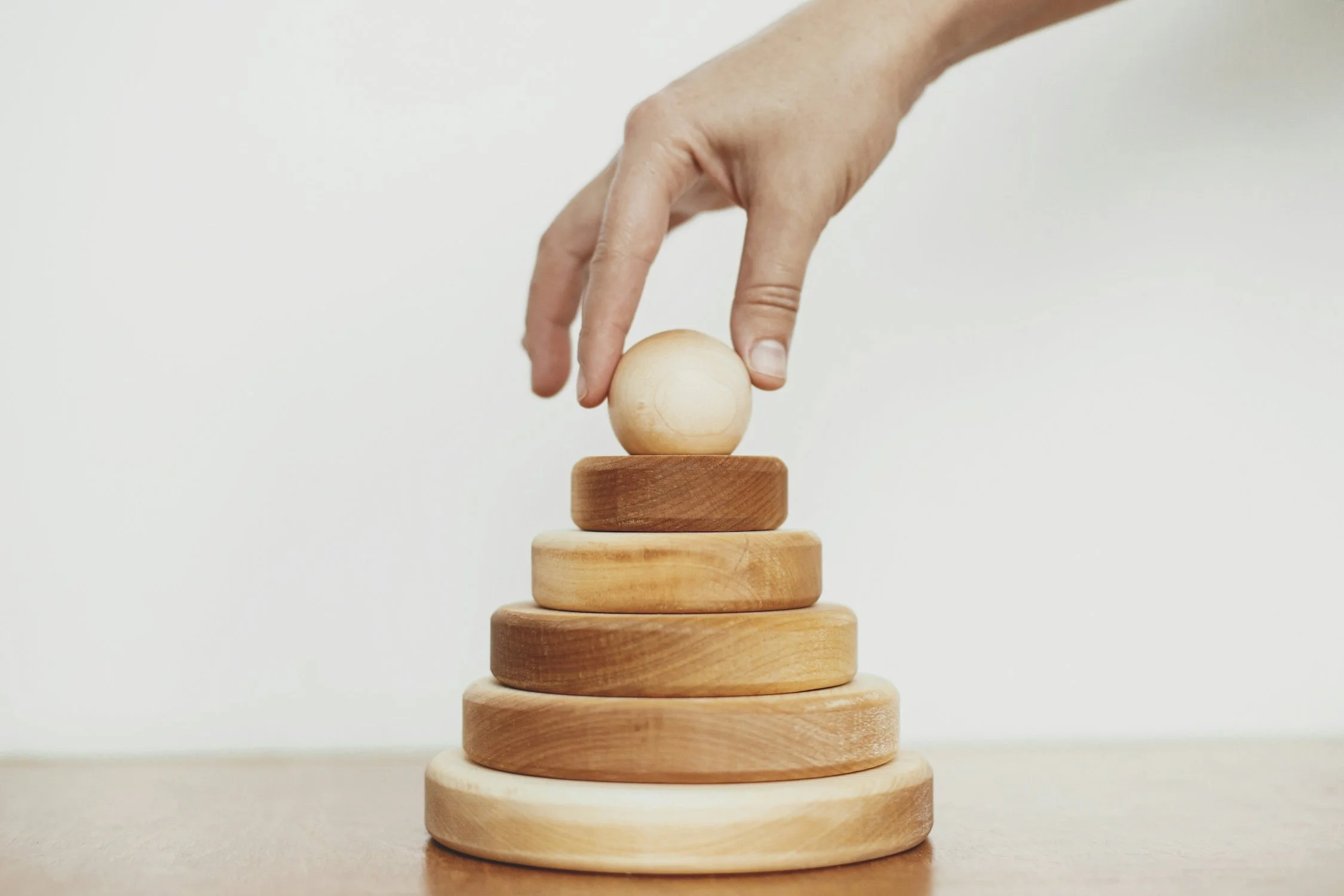 A person's hand placing a wooden sphere on top of a stack of balanced wooden rings, symbolizing the deliberate act of setting and aligning leadership priorities.