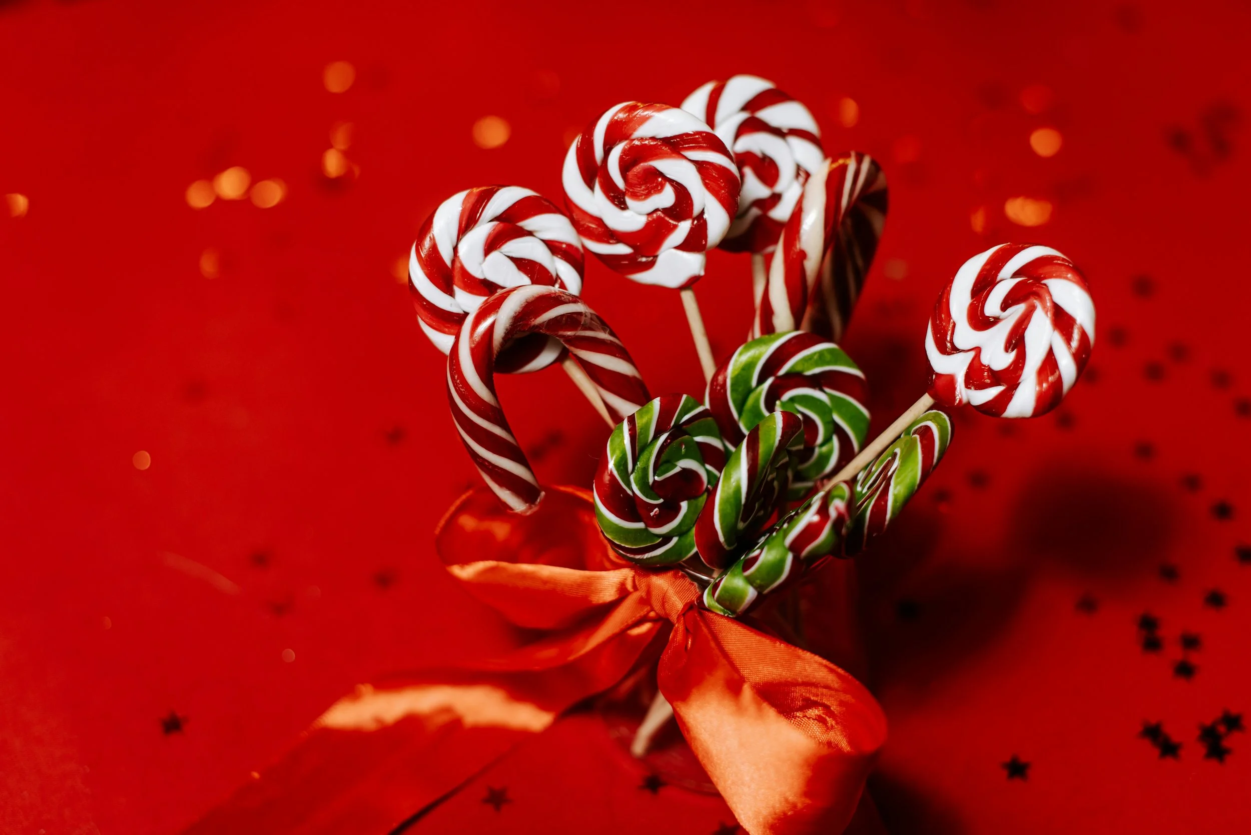 A festive bouquet of red, white, and green swirl lollipops and candy canes wrapped in an orange ribbon on a red background with star confetti.