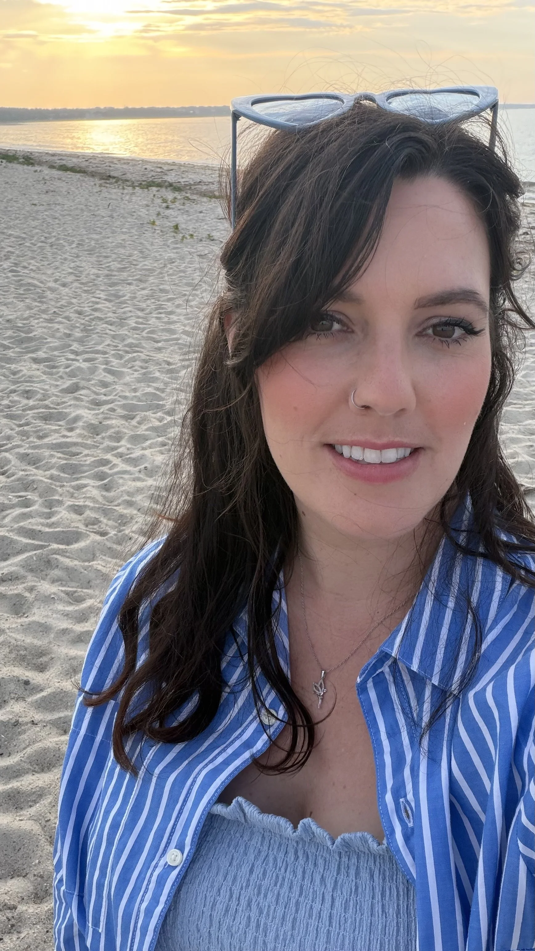 A woman with long brown hair, wearing sunglasses on her head, smiling at the camera, standing on a sandy beach during sunset, with water and sky in the background.