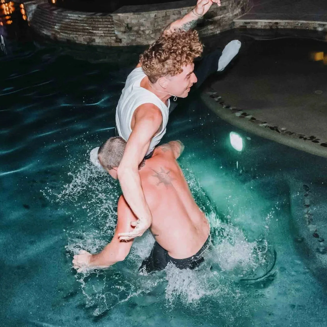 Two men wrestling in a swimming pool, one man with curly hair and the other with short blonde hair, with one gripping the other from behind.