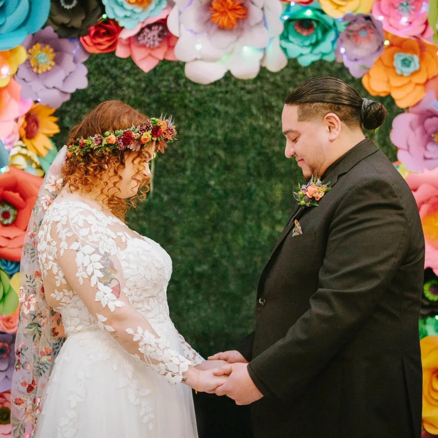 A bride and groom holding hands during their wedding ceremony, standing in front of a colorful floral backdrop with a green hedge.